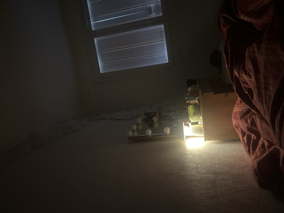 An interior scene depicts a dimly lit room, identified as a bedroom, located in Hauts de Bienne, France. In the foreground, a rumpled red and orange striped duvet covers a bed. To the right, a light-colored wooden nightstand is present, upon which a metal desk lamp with a white shade emits a bright, warm yellow light onto the adjacent wall and objects. Several small objects, including bottles and a green container, are arranged on the nightstand's surface. Above the nightstand, a wall-mounted vertical fixture featuring multiple spherical light bulbs is visible, with some shelves below it holding additional indistinct items. The left wall exhibits a textured pattern and a significant, irregular light-colored vertical streak, resembling peeling material or damage, extending from the ceiling down the corner where the left wall meets the back wall. On the right side of the frame, a large, two-paneled white-framed window is covered by vertical blinds or translucent curtains, allowing faint vertical streaks of external light to penetrate. No individuals are present in the scene.