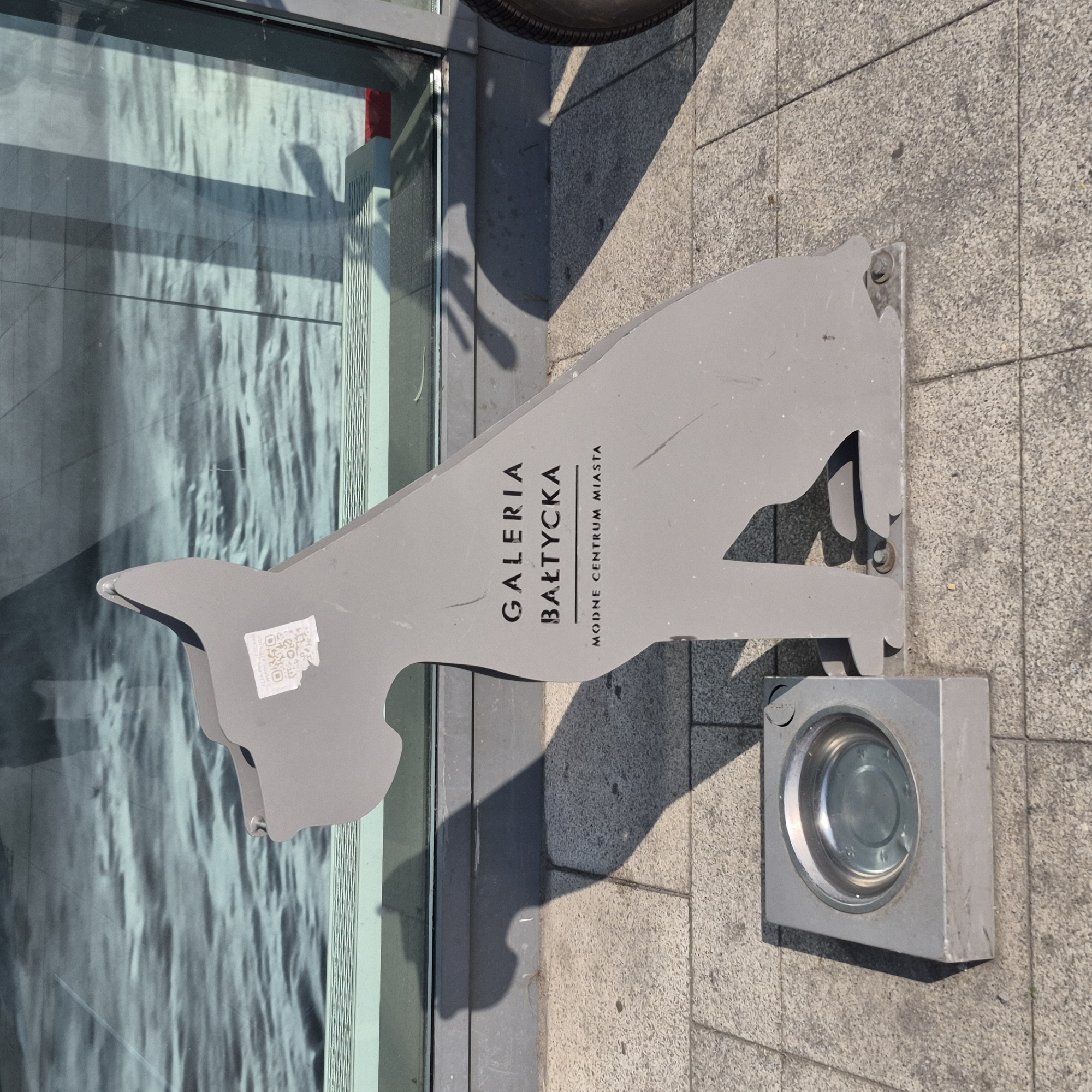 The image shows a metal sculpture of a dog's silhouette placed on a tiled sidewalk. The sculpture is gray and has text on it, reading "GALERIA BAŁTYCKA" and "MODNE CENTRUM MIASTA". Adjacent to the sculpture is a metal dog water bowl, which appears to be partially filled with water. In the background, a large window reflects a body of water with gentle waves. The lighting suggests it is daytime with sunlight casting shadows on the ground. The sculpture is located in the city of Gdańsk, Poland.