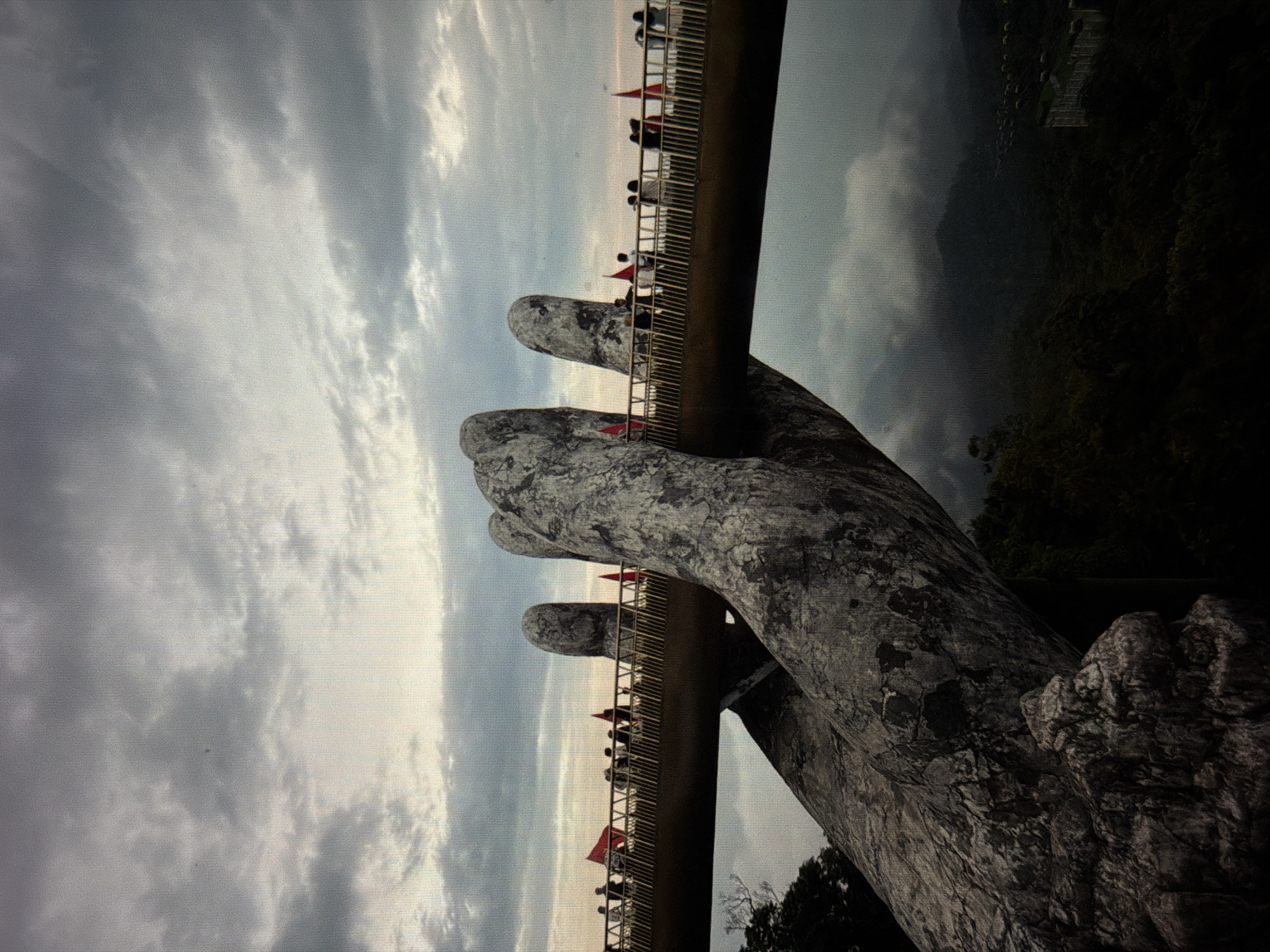 The image shows the Golden Bridge in Đà Nẵng, Vietnam. The bridge, supported by two giant stone hands, is filled with people walking along it. Red flags are visible on the side of the bridge. The sky is cloudy, and the background features mountains and vegetation. The hands supporting the bridge are textured to resemble ancient stone.