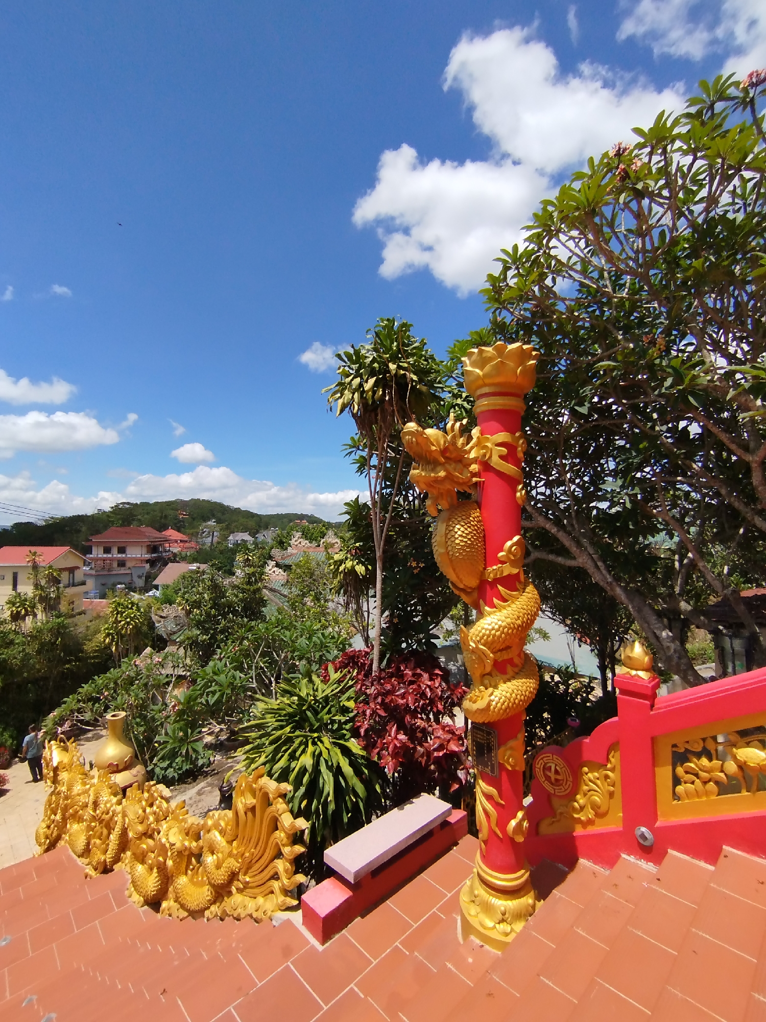 This image showcases a vibrant outdoor scene likely in Da Lat, Vietnam, considering the cultural elements. The focal point is a red column embellished with golden dragon sculptures, indicative of Southeast Asian temple architecture. The stairs and ornate golden railings suggest a pathway or a temple complex. Lush greenery, including trees and various plants, surrounds the structures. In the background, a cityscape with houses and buildings nestled amidst rolling hills can be observed under a blue sky with scattered white clouds. In the lower left a person stands on the steps.