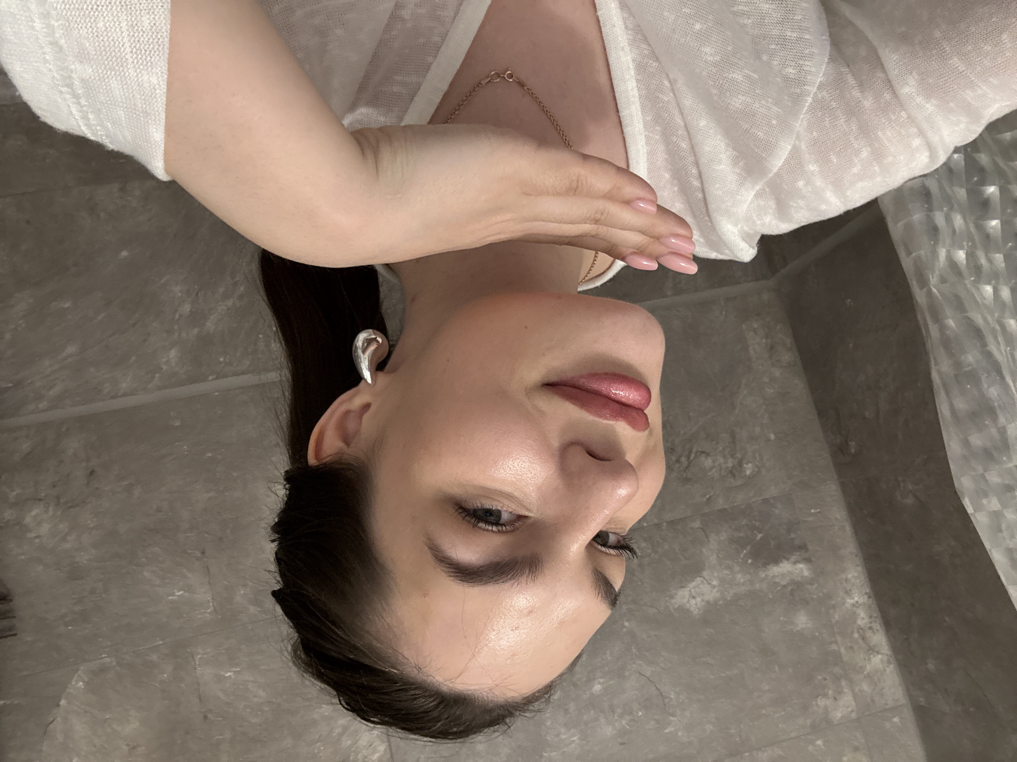 A woman with long, dark hair pulled back in a ponytail is positioned at an unusual angle, appearing to be upside down. She is wearing a white top and a delicate gold necklace. The nails on her hand, which is raised towards her face, are manicured with a light pink polish. She has an earring in her left ear. The background appears to be a tiled wall or floor with a gray, stone-like texture. The location is not discernible from the provided image. The city of Chaikovsky, Russia, cannot be confirmed as the location.