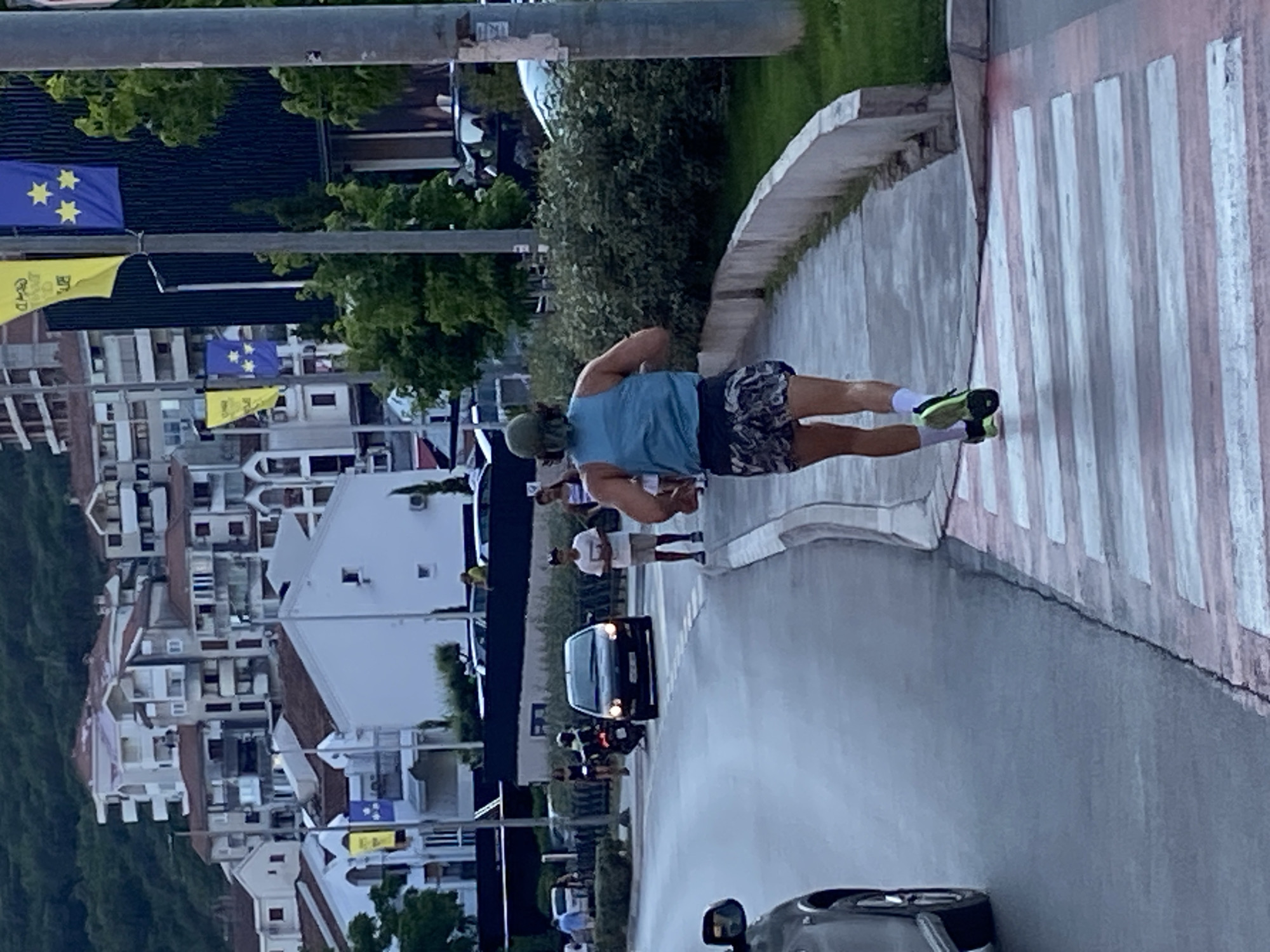 A person is jogging across a pedestrian crossing on a street in Budva, Montenegro. The individual is wearing a green cap, a blue tank top, and patterned shorts. They are also wearing white socks and green and black running shoes. The pedestrian crossing has alternating white and pink stripes. In the background, there are buildings with red tile roofs, trees, and flags. A car is visible on the left side of the image, and other people and a motorcycle are further down the street. The sky appears overcast.