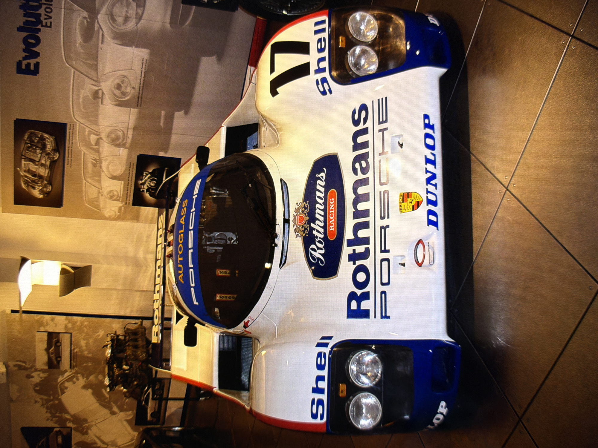 The photograph shows a white and blue Porsche 962 race car with various sponsor decals, including "Shell," "Rothmans," "Porsche," and "Dunlop." The car is displayed indoors, possibly in a museum or exhibition. The background features a wall with a large graphic depicting a series of cars and the word "Evolution." There are also images of a car engine and a schematic of a car. The lighting is dim, with a bright light source illuminating the car from above. The floor is tiled with a diamond pattern. The scene appears to be a static display of automotive history. The context of this photograph being located in Kyiv, Ukraine, is not evident from the visual information provided.