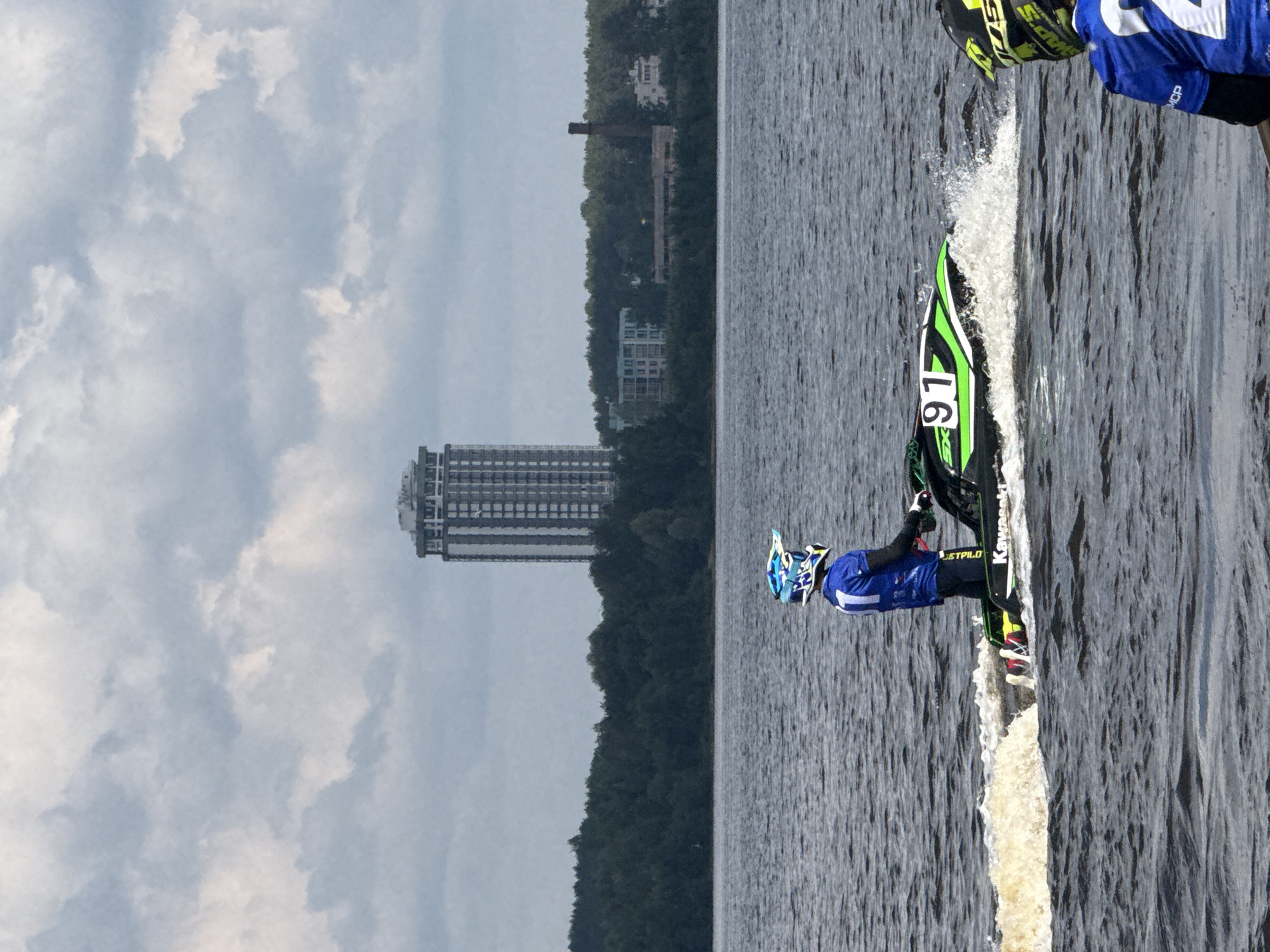 An aerial shot captures a jet ski race on the Kama River in Perm, Russia, on a cloudy day. The foreground features a jet skier in a blue and white helmet and a blue racing suit, identified by the number "1" on their back, riding a green and black jet ski with the number "91" displayed prominently. The jet ski is creating a spray of water as it moves. In the mid-ground, a dense line of dark green trees borders the river, beyond which several buildings are visible. A tall, modern skyscraper with a white facade and numerous windows stands out amongst these structures. The background is dominated by a cloudy sky. Another jet skier, wearing a yellow and black helmet and a blue racing suit with the number "2" visible, is partially visible in the lower right corner of the frame, looking towards the race. The overall scene suggests an outdoor sporting event taking place on a body of water with an urban backdrop.