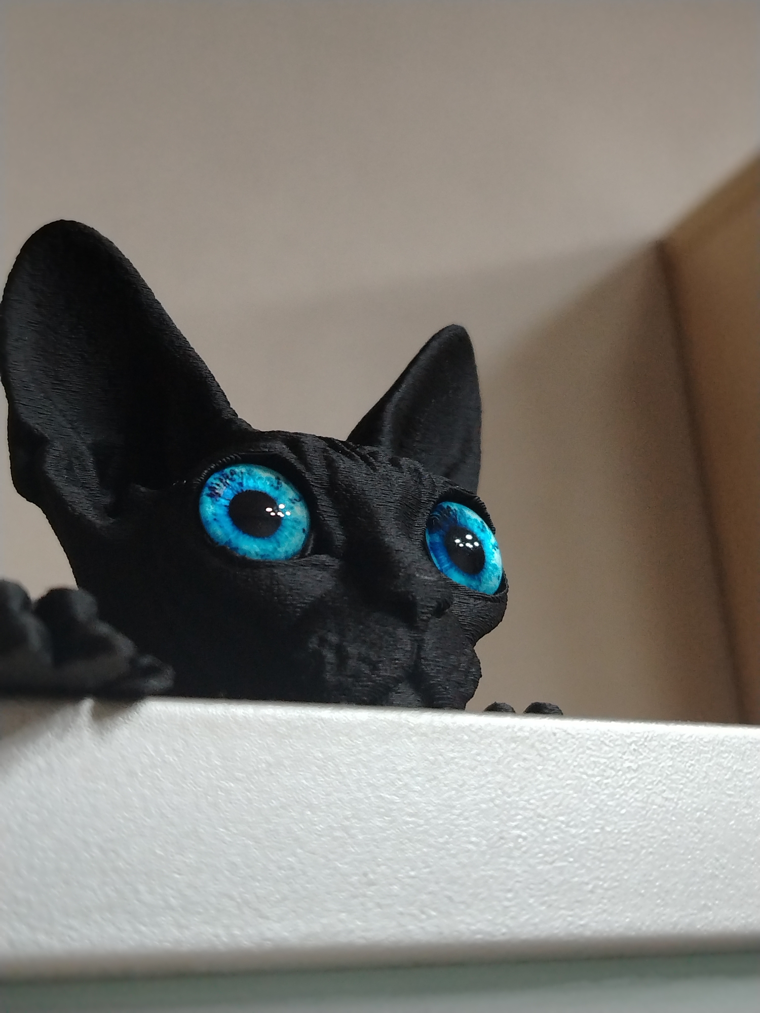 A black cat figurine with striking blue eyes peers over the edge of a white surface. The cat's ears are pointed, and its texture appears slightly rough, possibly indicating it's a 3D-printed object. The background is a blurry beige and white. There is no indication of the location of Izhevsk, Russia.