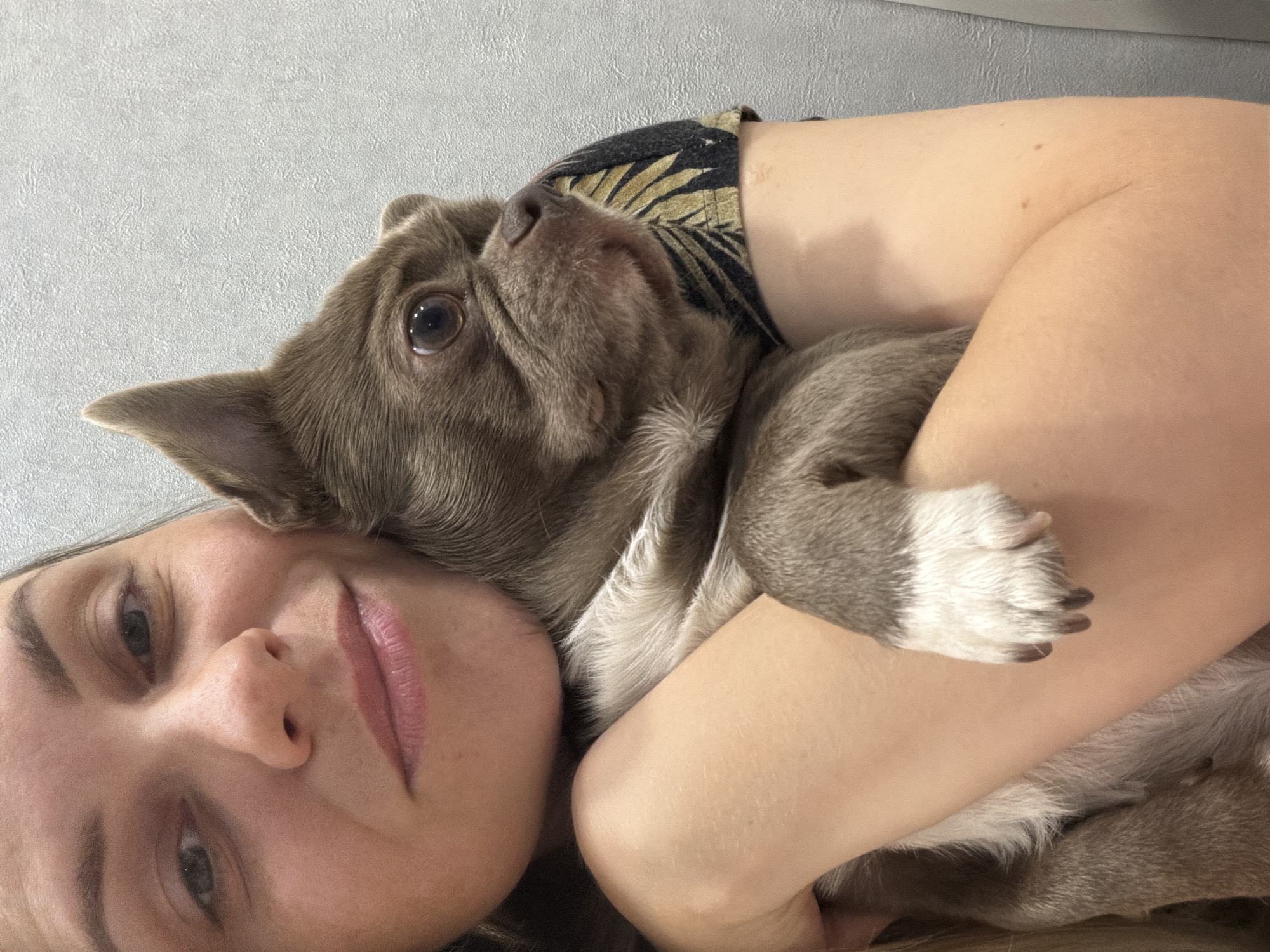 A selfie-style photograph shows a woman lying down, with a brown and white French bulldog cradled in her arms. The woman's face is close to the camera, and her eyes are open. Her lips are pink and slightly parted. The dog is snuggled against her chest, looking upwards. The dog's paws are visible, resting on the woman's arm. The background is a light gray wall. No specific landmarks or details indicate that this image was taken in Votkinsk, Russia.