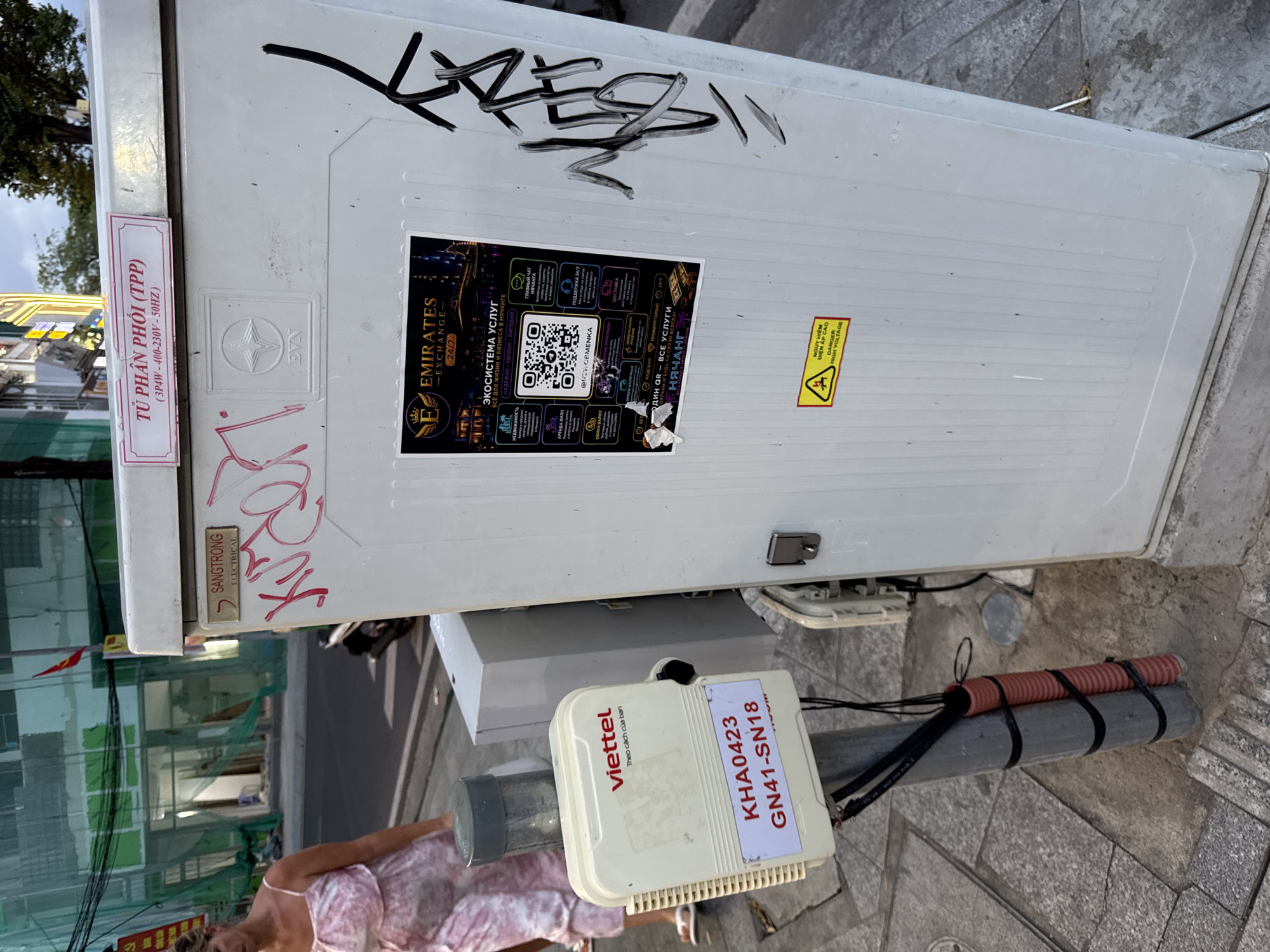 The image captures a street scene in Nha Trang, Vietnam, featuring utility boxes and infrastructure. In the foreground, a white Viettel box is mounted on a gray pole, displaying the label "KHA0423 GN41-SN18". Red and black wires are bundled and secured to the pole with black tape. To the right, a larger, white utility cabinet with vertical ridges is visible. This cabinet has a sticker featuring a QR code and text in Russian and English, advertising "EMIRATES EXCHANGE - 24/7" and various services. Above this sticker, there's a metallic plate with the text "SANGTRONG ELECTRICAL" and some pink graffiti. Another label on the top of the cabinet reads "TƯ PHÂN PHỐI (TPP)" with "(3P4W - 400-230V - 50HZ)" below it. A yellow triangular warning sticker with a lightning bolt symbol and text in Vietnamese and English ("NGUY HIỂM ĐIỆN ÁP CAO", "DANGER HIGH VOLTAGE") is affixed to the lower right section of the cabinet. In the background, a woman in a pink dress is walking on the sidewalk, and a building under construction, partially covered with green netting, is visible. The ground is paved with light-colored square tiles. The lighting suggests it is daytime, with diffused light and shadows present.