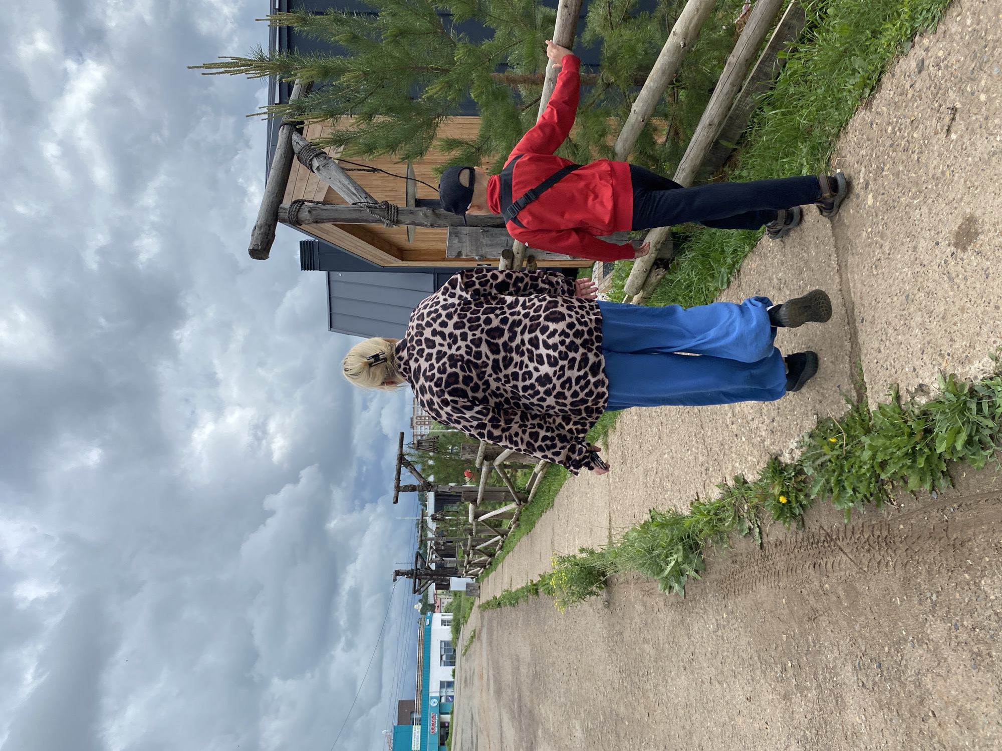 In Votkinsk, Russia, under a cloudy sky, two individuals walk away from the camera on a concrete path. The person in front, wearing a leopard-print jacket and bright blue pants, walks on the left side of the path. To their right, a younger person, in a red jacket and dark pants, walks slightly ahead, touching a wooden fence. The path is bordered by grass and wildflowers. Behind the fence, a modern building with wooden accents and a dark roof is visible, along with a pine tree. In the background, a street with buildings and power lines suggests an urban or suburban setting.