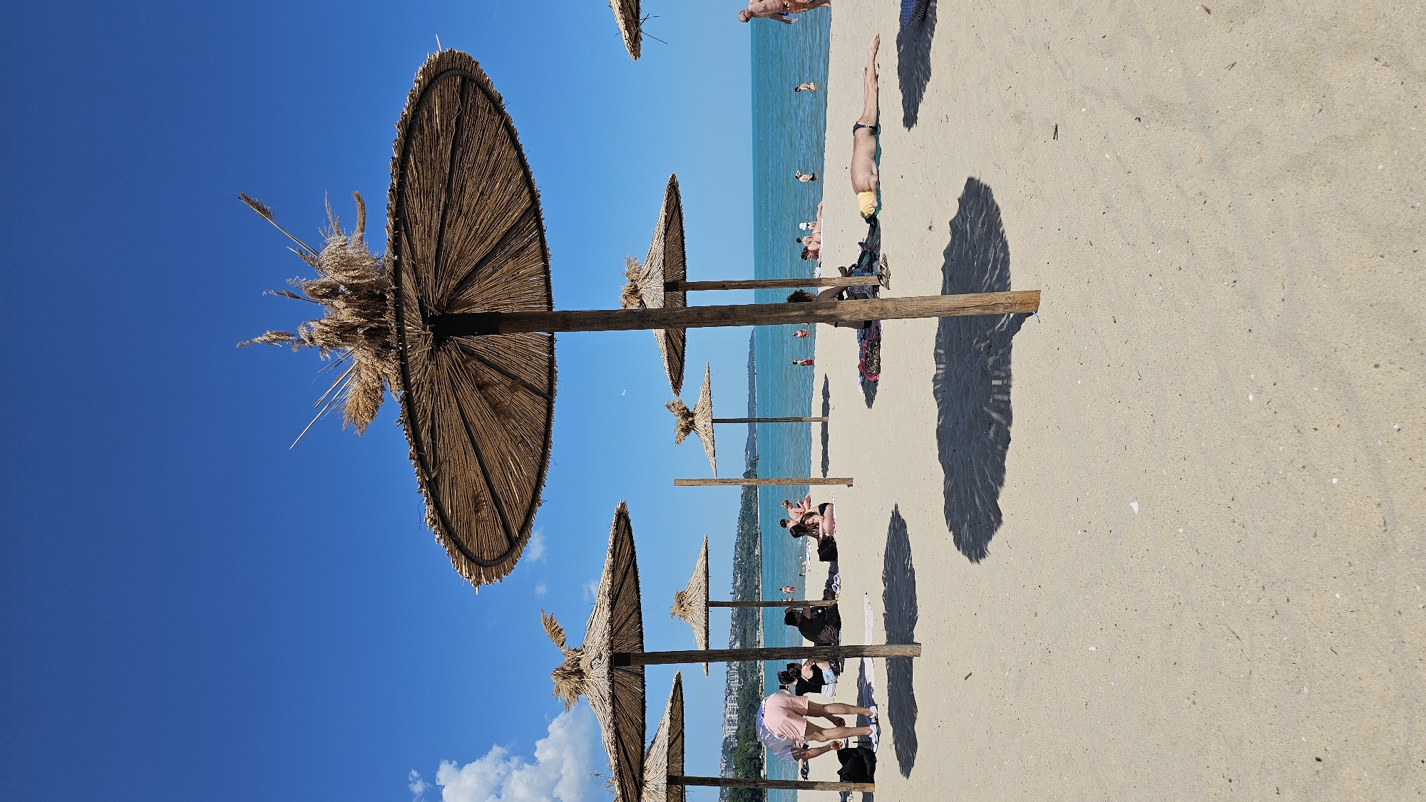 The image shows a beach in Varna, Bulgaria, on a sunny day. The scene is dominated by rows of straw umbrellas providing shade for beachgoers. The bright blue sky is partially visible between the umbrellas. People are seen lounging on the sand, swimming in the sea, and walking along the shore. In the background, buildings are visible, indicating the proximity of the city. The shadows of the umbrellas are cast on the sand.