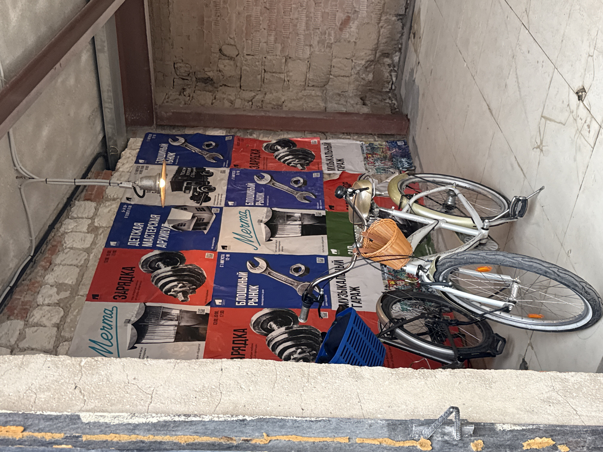 An indoor scene features two bicycles parked against a brick wall adorned with posters. The posters are in various colors and sizes, depicting graphics of dumbbells, wrenches, trucks, and architectural drawings, with Cyrillic text, indicating events or advertisements. One bicycle is silver with a wicker basket on the handlebars and a small basket on the rear rack. The other bicycle is dark-colored and partially obscured by the silver one, with a blue plastic basket attached to its handlebars. The environment appears to be a covered outdoor space or a garage, with exposed brick walls, a concrete floor, and overhead electrical conduits and a light fixture. The scene suggests a creative or industrial space, possibly in Vnukovo, Russia, given the context.