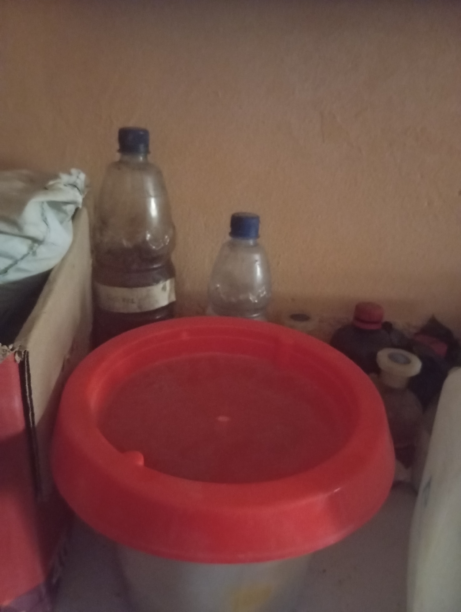 The image shows a collection of items on a shelf against a textured, peach-colored wall. In the foreground, a large, bright red, circular basin or lid is prominently displayed. Behind it, to the left, is a tall, clear plastic bottle with a blue cap, containing a dark liquid and a label. To the right of this bottle, a smaller, clear plastic bottle with a blue cap is visible. Further to the right, there are several other objects including a bottle with a red cap, and a white object that could be a bottle or container. A cardboard box is partially visible on the far left. The scene appears to be an indoor storage area, possibly a shelf in a home or shop, located in the city of Jalingo, Nigeria.