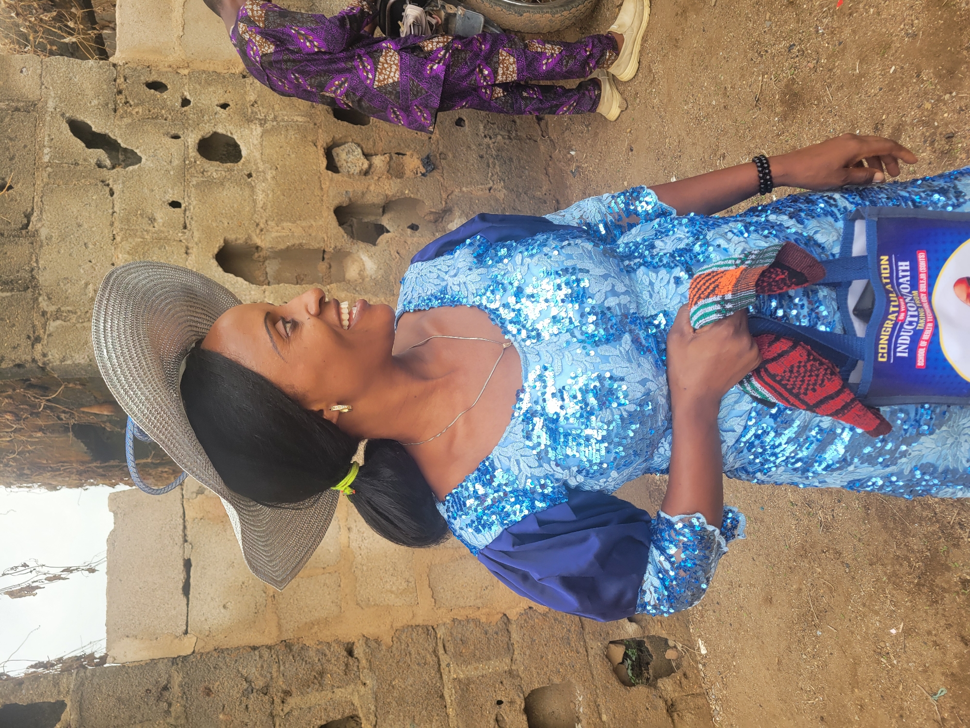 A woman in a light blue sequined dress and a wide-brimmed straw hat is standing outside in Madalla, Nigeria. She is holding a blue tote bag labeled "CONGRATULATIONS ON YOUR INDUCTION/OATH TAKING FROM SCHOOL OF HEALTH TECHNOLOGY GULEJA (SOHITE)." She is smiling and looking to her left. In the background, a person wearing a purple patterned outfit and white shoes is visible. The surrounding environment includes a brick structure and dirt ground.