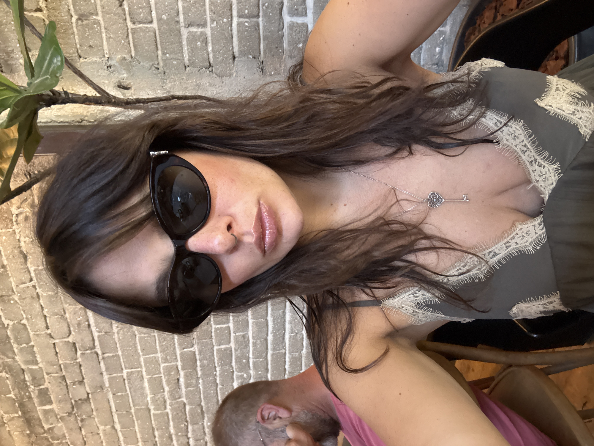 The image shows a selfie of a woman with long brown hair and sunglasses, wearing a dark green camisole with lace trim and a key-shaped necklace. She is sitting in front of a brick wall, with a plant to her right. In the background, a man with a beard and glasses is visible. The scene appears to be outdoors, possibly at a cafe or restaurant. The lighting suggests it is daytime. There are no specific visual cues in the image that would definitively place the location in Tel-Aviv, Israel.