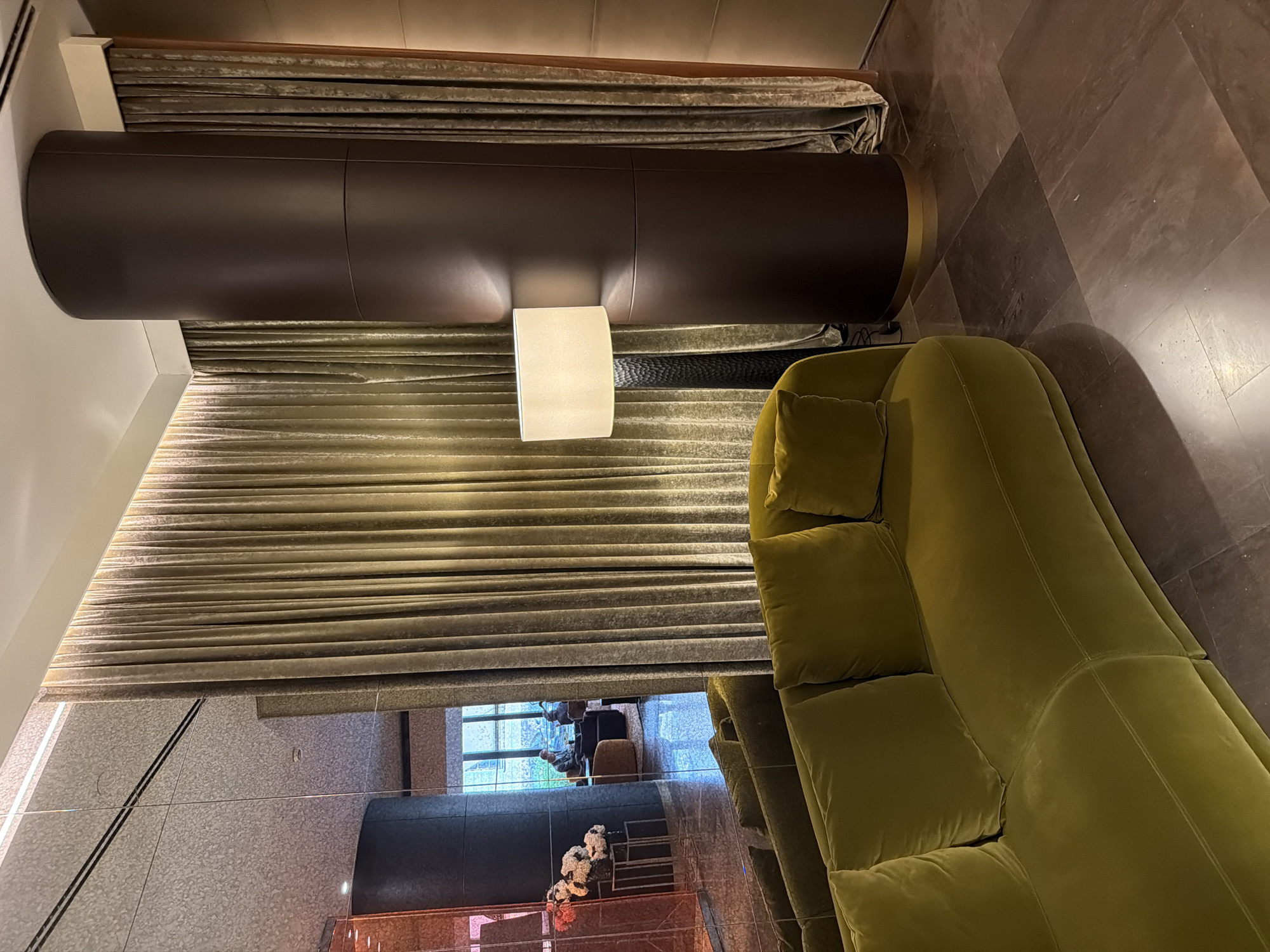 This image captures an interior scene featuring a plush, olive-green sofa in the foreground. Behind the sofa, a tall, dark brown cylindrical column stands prominently, draped with textured, golden-brown curtains. To the left of the column, a floor lamp with a white lampshade casts a warm glow. The background includes a large mirrored wall reflecting a seating area where two individuals are visible, one seated and looking at a laptop, the other in the distance. The floor is tiled with large, dark brown slabs. The environment appears to be a hotel lobby or a similar upscale public space, possibly in Saint Petersburg, Russia, given the context provided.