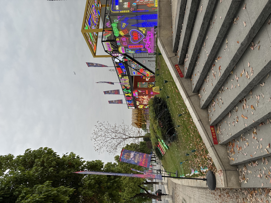 The image shows a set of stone steps leading up to a brightly lit building adorned with neon signs. The scene is located at 29.608° N, 106.588° E in Chongqing, China. The steps are covered in fallen leaves. The building appears to be a commercial establishment decorated with a variety of colorful neon signs, creating a vibrant visual display. There is a small, gray, single-door car parked on the street below, and a small plane is visible in the sky. The environment is characterized by a mix of urban and natural elements, with buildings, trees, and fallen leaves all contributing to the scene. There are no people visible in the image.