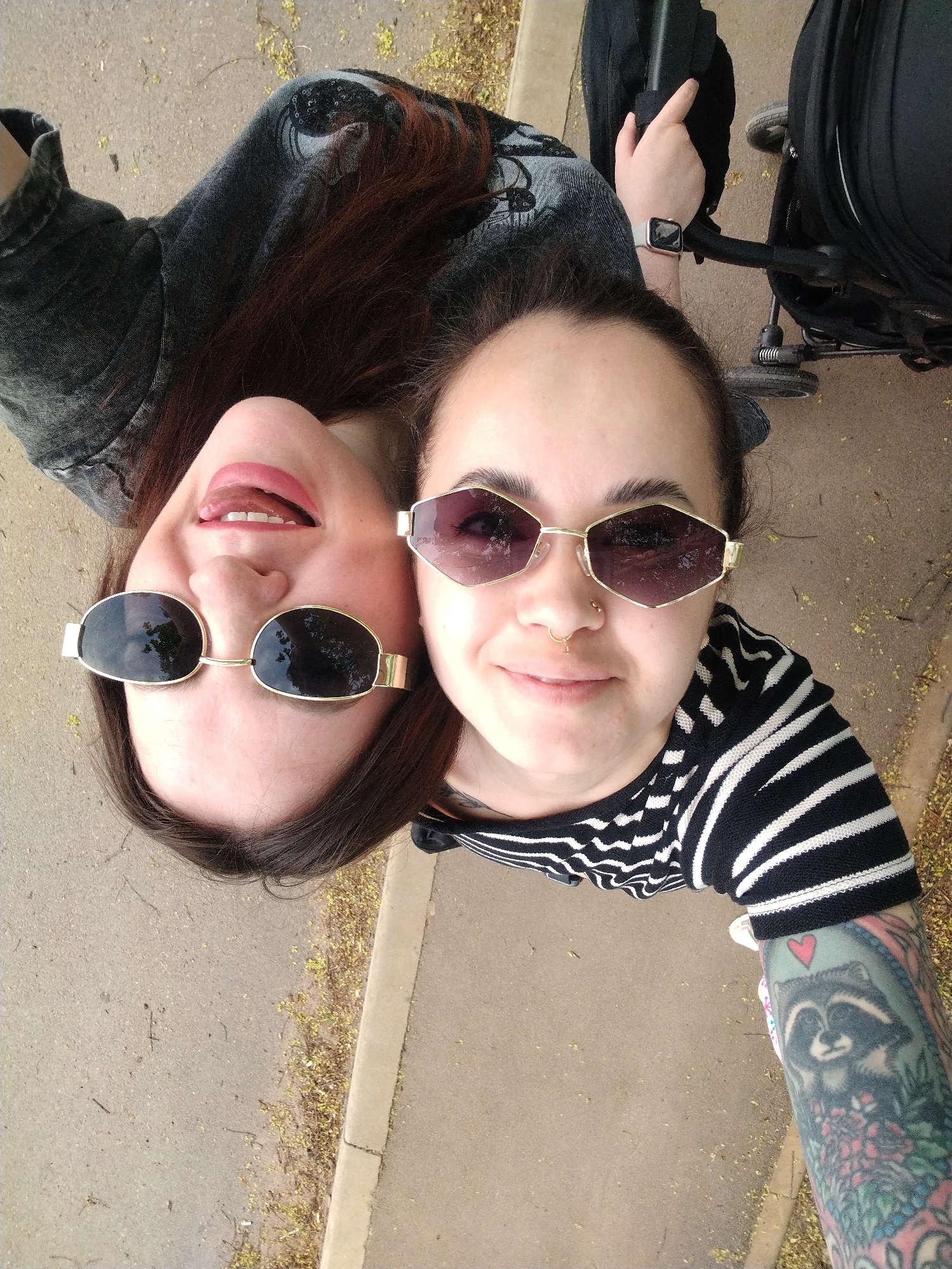 Two women are posing for a selfie outdoors. One woman wears sunglasses with rounded lenses and a dark denim jacket, with her mouth slightly open, tongue visible. The other woman is wearing geometric sunglasses, a striped shirt, and has a tattoo of a raccoon with a heart above its head on her arm. Both are fair-skinned. A stroller is partially visible in the background. The environment appears to be a paved outdoor area with scattered yellow debris, possibly fallen flowers or leaves. The location cannot be determined to be Noviy, Russia based on the image alone.
