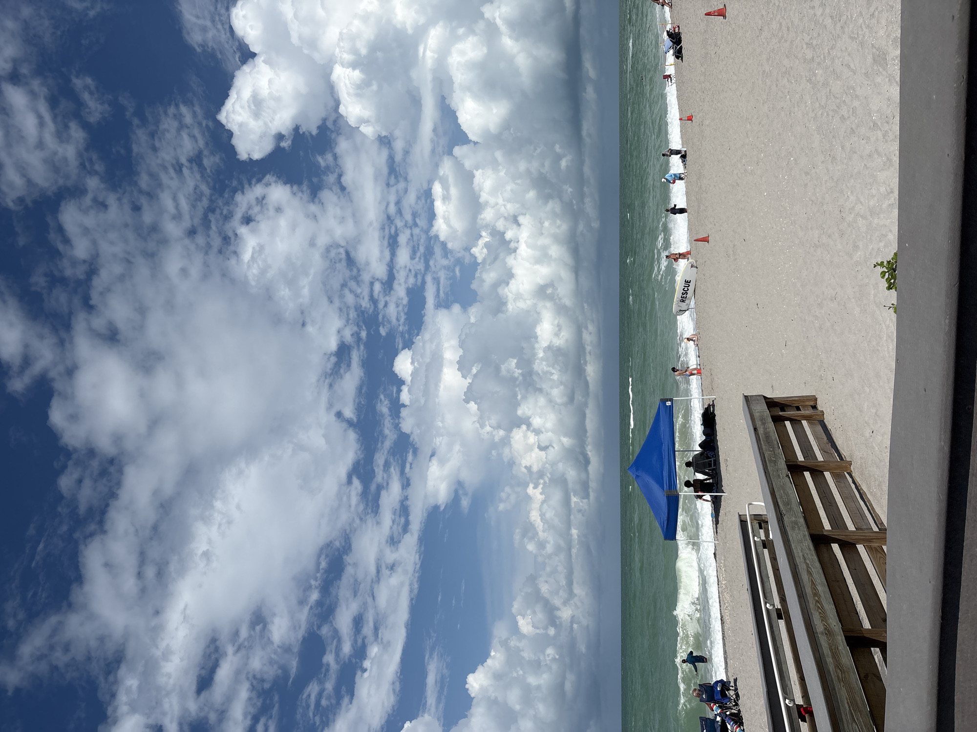 The image shows a beach scene in Englewood, United States. A wooden walkway leads down to the sandy beach. Several people are visible on the beach, some are sitting under a blue canopy, while others are near the water. The ocean has moderately sized waves and the water is a greenish hue. The sky is partly cloudy with large, white cumulus clouds dominating the upper portion of the frame. A rescue board is propped up on the sand near the water's edge. Orange traffic cones are placed sporadically on the beach.