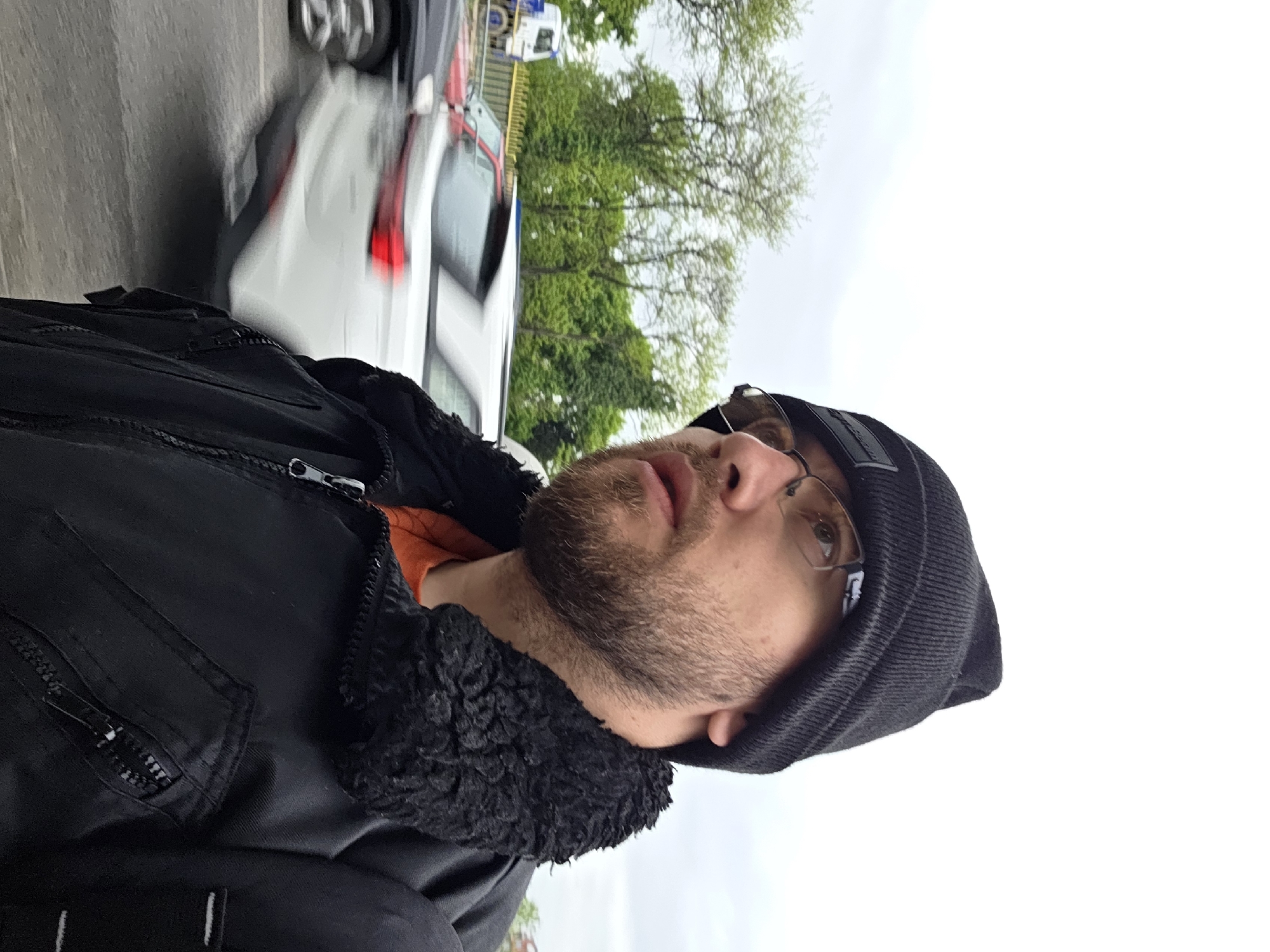 An eye-level portrait captures a man with a light brown beard and mustache, wearing glasses, a dark grey beanie, and a black jacket with a black faux fur or sherpa collar. An orange shirt is visible underneath his jacket. He looks slightly upwards and to his right, with a neutral expression and his mouth slightly ajar. The background features a blurred white car in motion on a road, indicating traffic, with green trees beyond it. The sky is overcast and bright. The scene is an outdoor, daytime setting, likely in an urban or semi-urban environment, with the provided context placing it in Plovdiv, Bulgaria. No discernible text is visible.