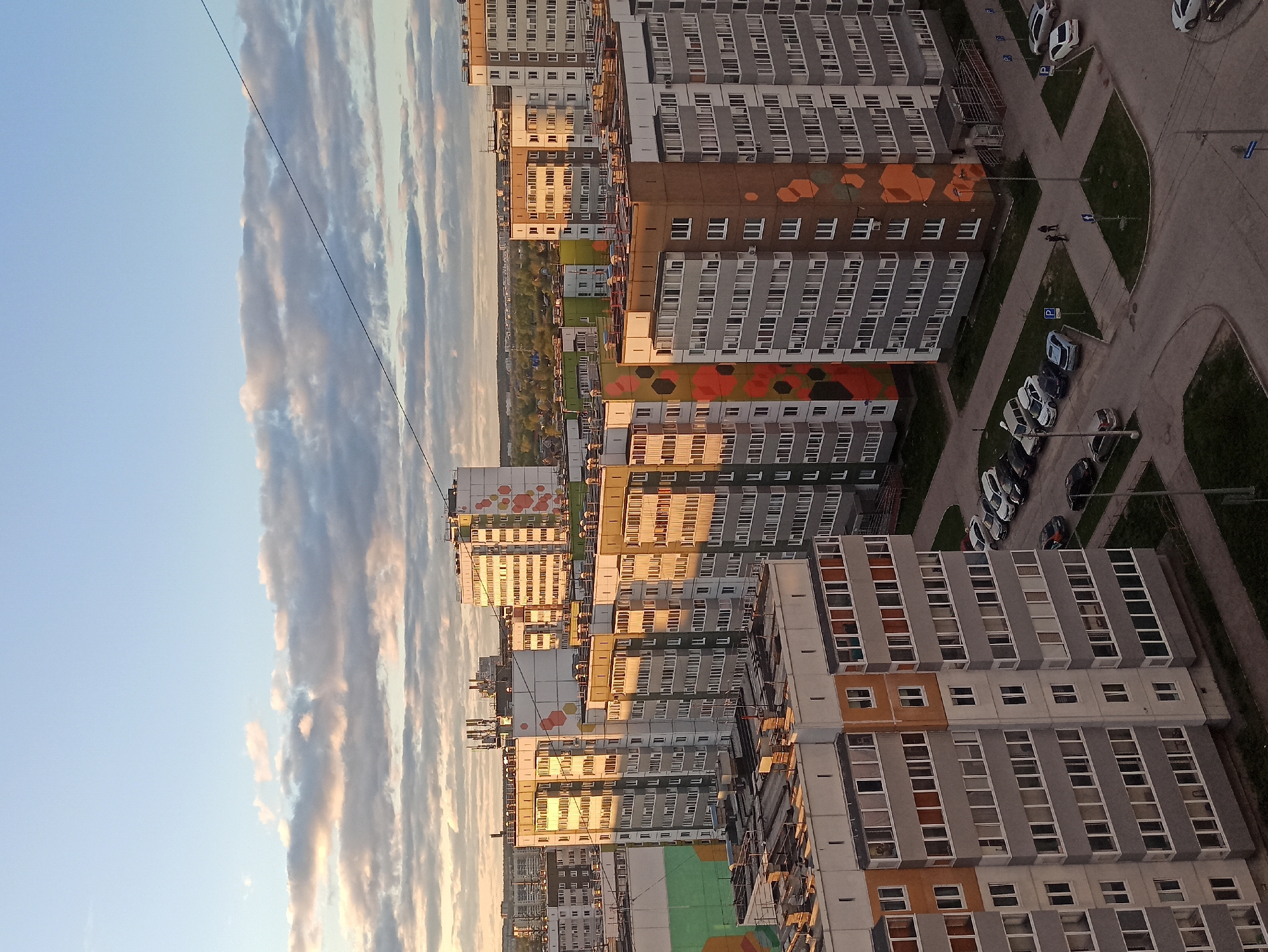 The image depicts a high-angle view of a residential area in Кондратово, Russia, under a sky with layered clouds during a sunset. Several apartment buildings with repetitive window patterns dominate the scene. Some buildings have colorful geometric patterns on their facades. Cars are parked in designated parking areas, and a couple of people are walking along a paved path. A grassy area is visible between the buildings. Overhead electrical wires are seen against the sky.