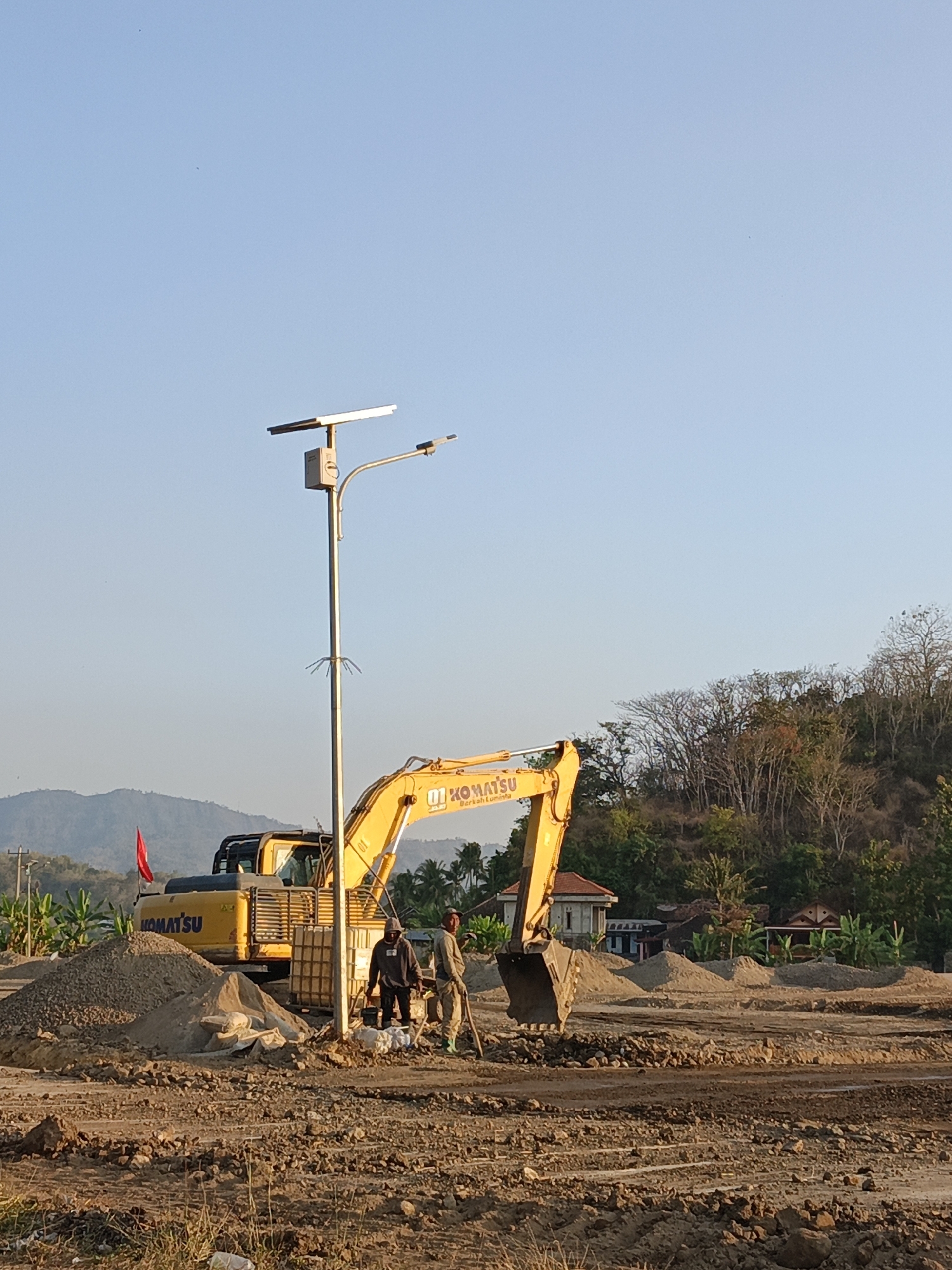 In the city of Krakitan, Indonesia, a construction site is depicted under a clear blue sky. The foreground shows disturbed earth and piles of gravel, indicating ongoing work. In the midground, a large yellow Komatsu excavator is positioned, its bucket resting on the ground. Two men, dressed in casual work attire, stand near the excavator. One is holding a shovel. Adjacent to them is a solar-powered street light pole with a rectangular panel at the top. Behind the excavator, more piles of sand and gravel are visible, along with some buildings and lush vegetation. In the background, rolling hills covered in trees rise, completing the rural landscape.