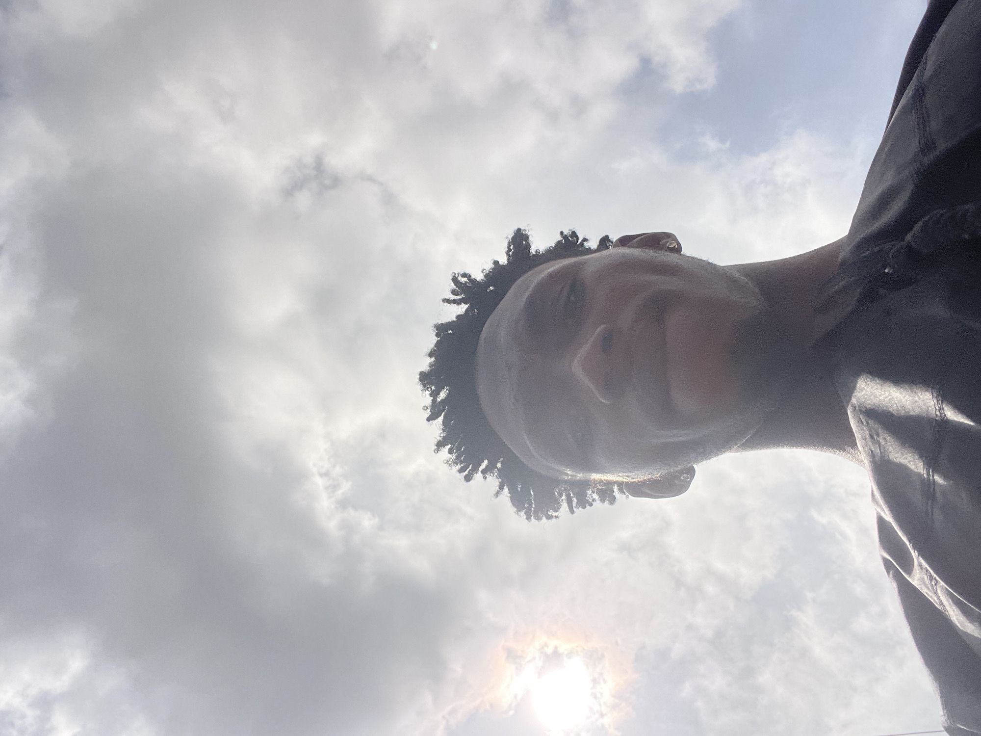 A low-angle, eye-level shot captures a person's head and upper torso against a bright, cloudy sky in Lokoja, Nigeria. The individual, with dark skin and dark, curly hair, is looking towards the camera with a subtle, serene expression. Their face is angled slightly upwards and to the left, with the right side of their face receiving more light. The sun, partially obscured by clouds, is visible in the bottom right, casting a bright flare and illuminating the edges of the person's hair and profile. The sky is predominantly filled with white and light grey clouds, indicating a partially overcast but bright daytime. The person is wearing dark-colored clothing, possibly a jacket or shirt, visible on the right side of the frame. No text is visible in the image.