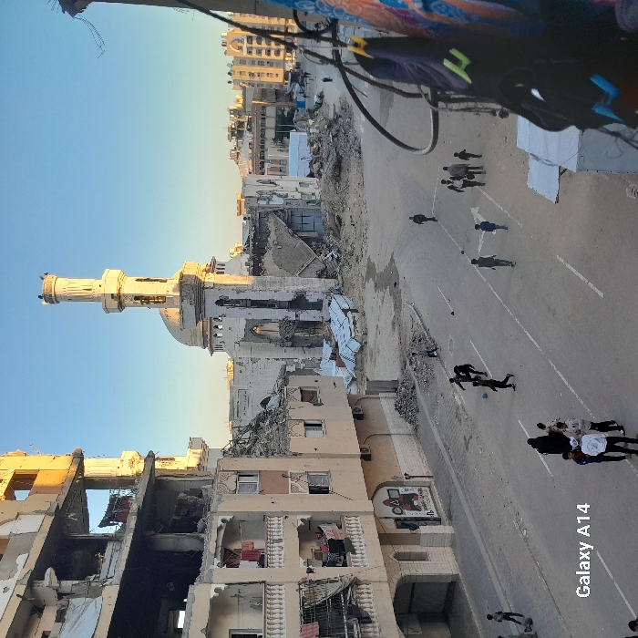 The image captures a street scene amidst war-torn buildings, with a mosque prominently standing partially destroyed. People walk on the street, suggesting movement and life persisting despite the surrounding devastation. The buildings on either side of the road are heavily damaged, exposing interiors and indicating the scale of destruction. The presence of a clear sky contrasts sharply with the ground-level chaos, creating a poignant juxtaposition.