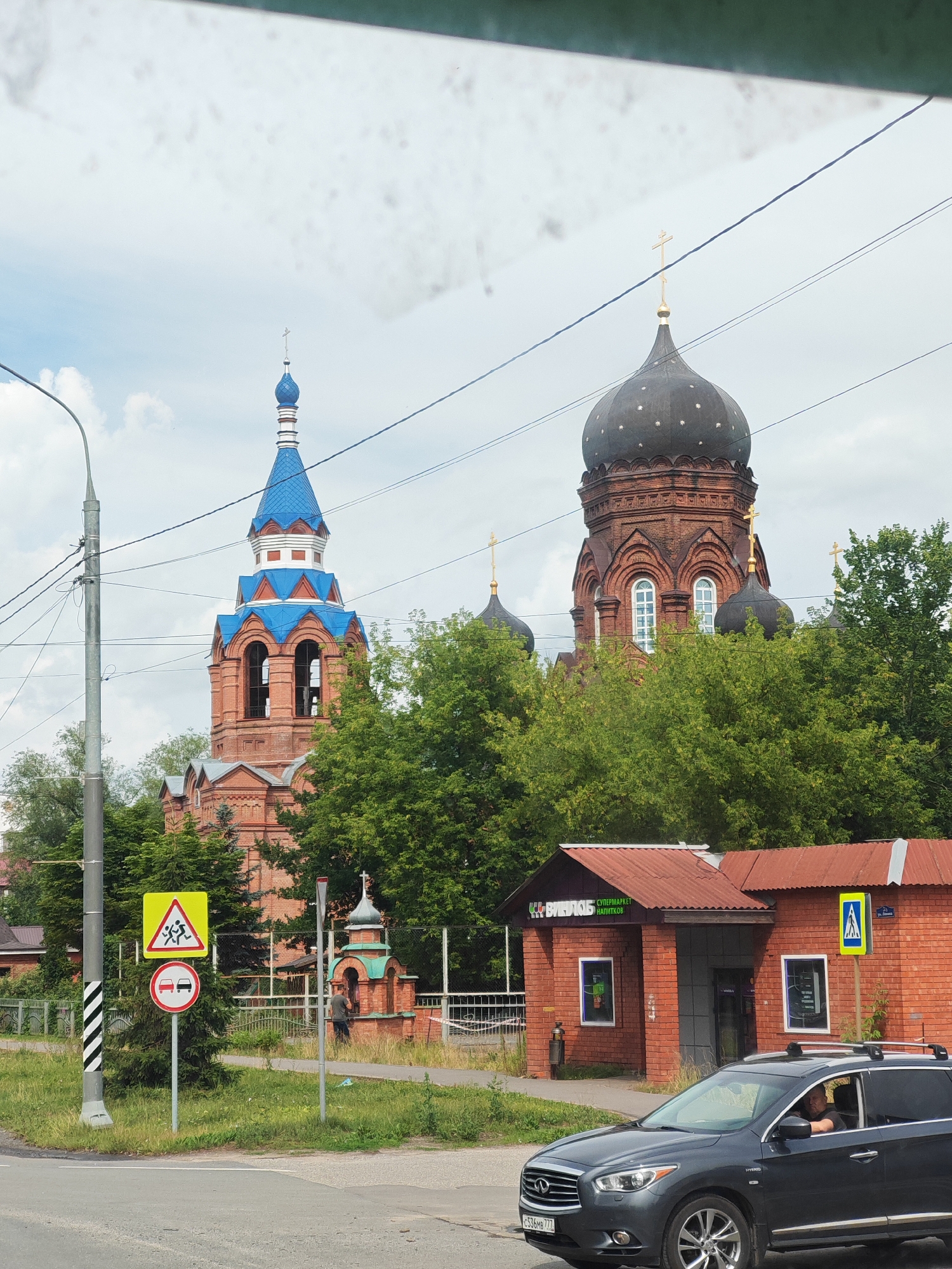 The image captures a street scene in Likino-Dulyovo, Russia. Two prominent brick churches with distinctive onion domes dominate the background. The church on the left has a blue-domed bell tower, while the larger church on the right features a dark, star-speckled dome. Both are adorned with gold crosses. The foreground shows a paved road with a dark gray SUV driving towards the right. A person is visible inside the SUV, looking towards the left. On the side of the road, there is a sidewalk, a grassy area, and several traffic signs. One sign, a yellow triangle with red border, depicts children running, indicating a school zone or playground. Another sign, a red circle with a horizontal line and two cars, signifies no passing. Adjacent to these signs is a small, green-domed brick chapel. A brick building with a red roof, identified as a supermarket by the text "СУПЕРМАРКЕТ НАПИТКОВ" (Supermarket of Drinks) and a logo with the word "ВИЛЮЙ", is located to the right of the chapel. Tall green trees are interspersed throughout the scene, framing the buildings and contributing to a lush, semi-urban environment. Overhead power lines are visible crisscrossing the sky. The weather appears overcast with scattered clouds.