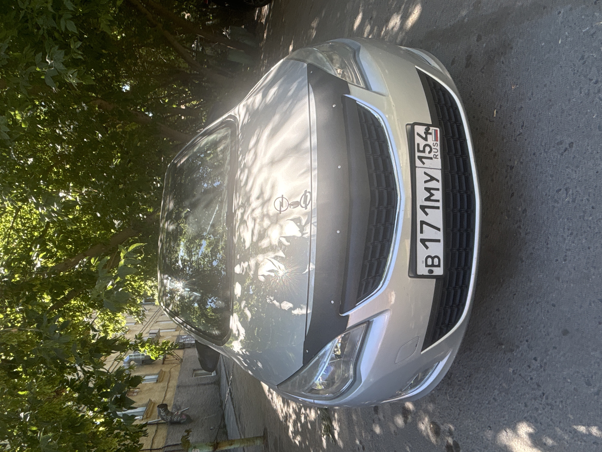 A silver Opel Astra is parked on an asphalt surface, possibly a street or parking lot, in front of a light-colored building with several windows. Lush green trees overhang the car, casting dappled shadows on its hood and the surrounding area. The car's license plate is visible and reads "B 171MY 154 RUS". The scene appears to be set in a residential area of Novosibirsk, Russia, on a sunny day. There are no people or animals visible in the frame.