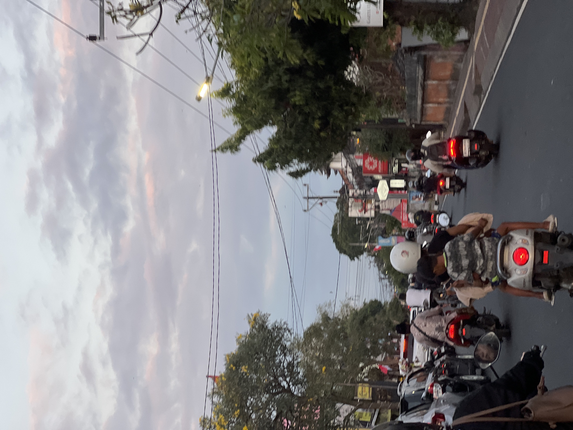 A traffic scene unfolds in Denpasar, Indonesia, under a sky painted with soft pastel clouds. Several motorbikes navigate a street lined with trees and buildings, some adorned with red and white flags. The road is a mix of asphalt and what appears to be a yellow painted lane.
In the foreground, a person on a motorbike is partially visible, their black jacket and brown bag suggesting a casual attire. Further ahead, a motorbike carries two individuals; the rider wears a white helmet, and a young child sits behind them, clad in a gray and white striped shirt. Red taillights from other motorbikes punctuate the scene, indicating the flow of traffic.
On the left side of the road, cars are visible, suggesting a busy urban environment. Trees, some bearing yellow flowers, cast shadows, and power lines crisscross overhead, illuminated by a street lamp. Buildings with traditional Balinese architectural elements, like patterned walls, are also present. The overall atmosphere suggests a late afternoon or early evening, with the fading light casting a gentle glow on the surroundings.