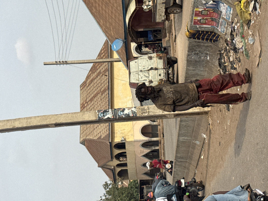 The image depicts a daytime street scene in Lafia, Nigeria, characterized by urban infrastructure and public activity.
In the foreground, a male individual stands facing slightly right, looking downward. He has dreadlocks and a full beard, appearing unkempt. He wears a dark, soiled short-sleeved t-shirt with obscured text, possibly reading "SASTODAY SAI JOS," and dirty, reddish-brown pants. His feet are bare or covered by minimal, worn footwear. To his left, a thick, grey concrete utility pole stands, featuring two political posters with black-and-white images of a man's head, one labeled "M. ENITO" and the other "M. MAGA."
The central figure is positioned on a dirt shoulder beside a paved road. A low concrete barrier separates this shoulder from the main roadway. Along the base of this barrier, to the right of the man, an accumulation of discarded refuse, including plastic, paper, and other debris, is visible. Two flattened posters, one displaying "STAR RADLER" branding, and a piece of striped fabric (blue and yellow/white) are also on the ground near the trash.
On the paved road to the left, two individuals on motorcycles are present. One rider wears a light blue shirt and blue jeans, while the other, wearing a red shirt and white cap, is seated on a dark motorcycle, facing away.
In the mid-ground, behind the concrete barrier and the central figure, a white, dilapidated panel van with visible rust and dirt is parked on a dirt patch. To the right of the van, a small, open-front establishment with a brown corrugated metal roof and a blue satellite dish is visible. A person is indistinctly seated inside this structure. Further right, a thinner utility pole with multiple power lines extends across the frame.
The background is dominated by a two-story building with yellow-painted walls and a multi-gabled brown corrugated metal roof. The ground floor features a series of arched openings. The sky above is light blue with a single small white cloud. Green foliage from trees is visible on the far left.