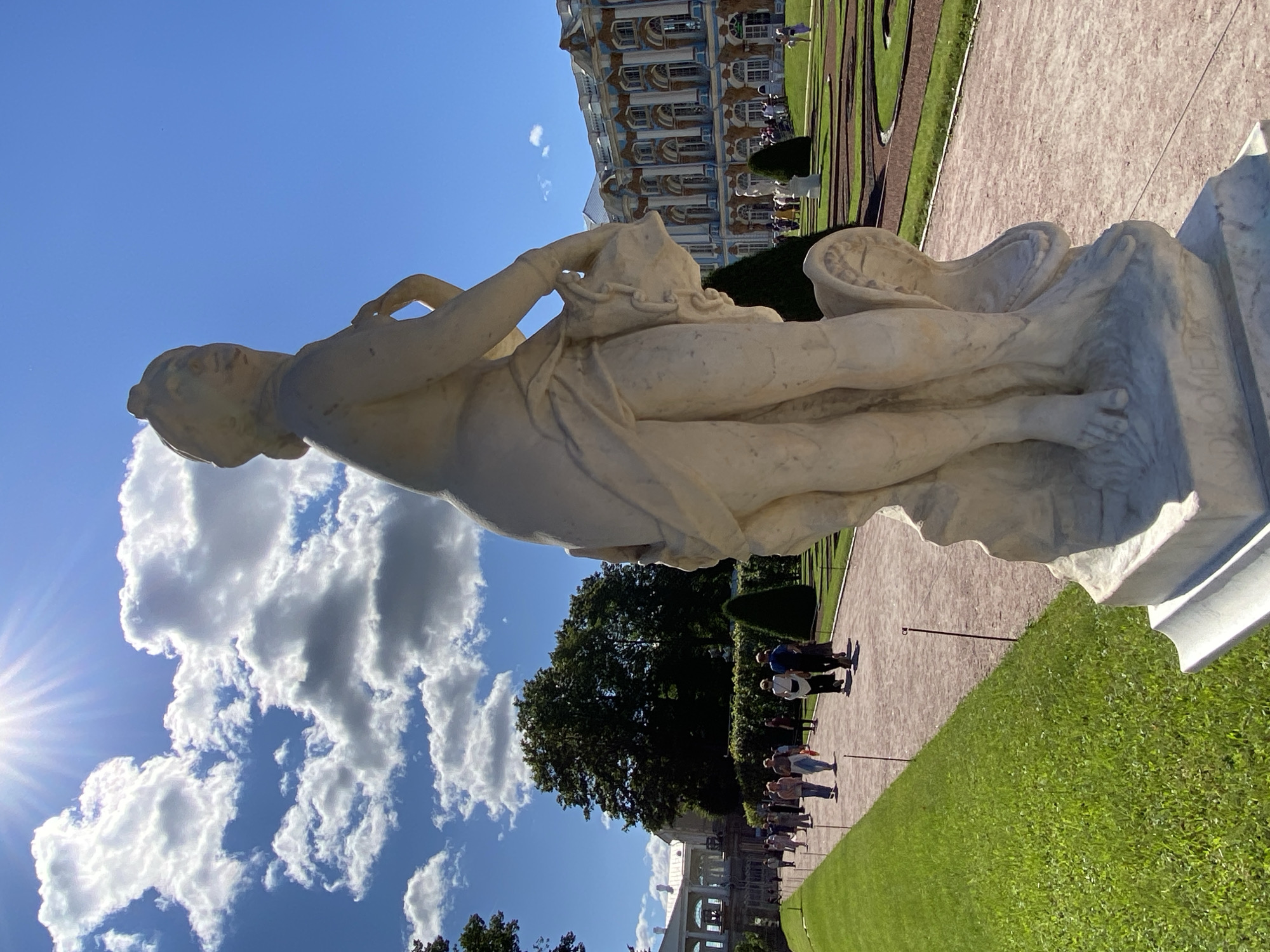 The image depicts a bright, sunny day in a park setting, likely in Pushkin, Russia, given the architectural style of the distant building. In the foreground, a weathered white marble statue of a female figure is prominently positioned, angled to the right. The statue appears to be partially draped, with one arm raised to her chest. The statue rests on a multi-tiered marble base. A pathway, made of reddish gravel, extends into the distance to the right of the statue. To the left of the pathway is a well-maintained green lawn. Further in the background, several people are walking along the pathway, dressed in casual attire. Behind them, a large, ornate building with a blue and white facade, featuring numerous arched windows and decorative elements, is visible. Several manicured evergreen trees and bushes are scattered throughout the park. The sky is a vibrant blue, with scattered fluffy white clouds and a bright sunbeam shining from the upper left corner.