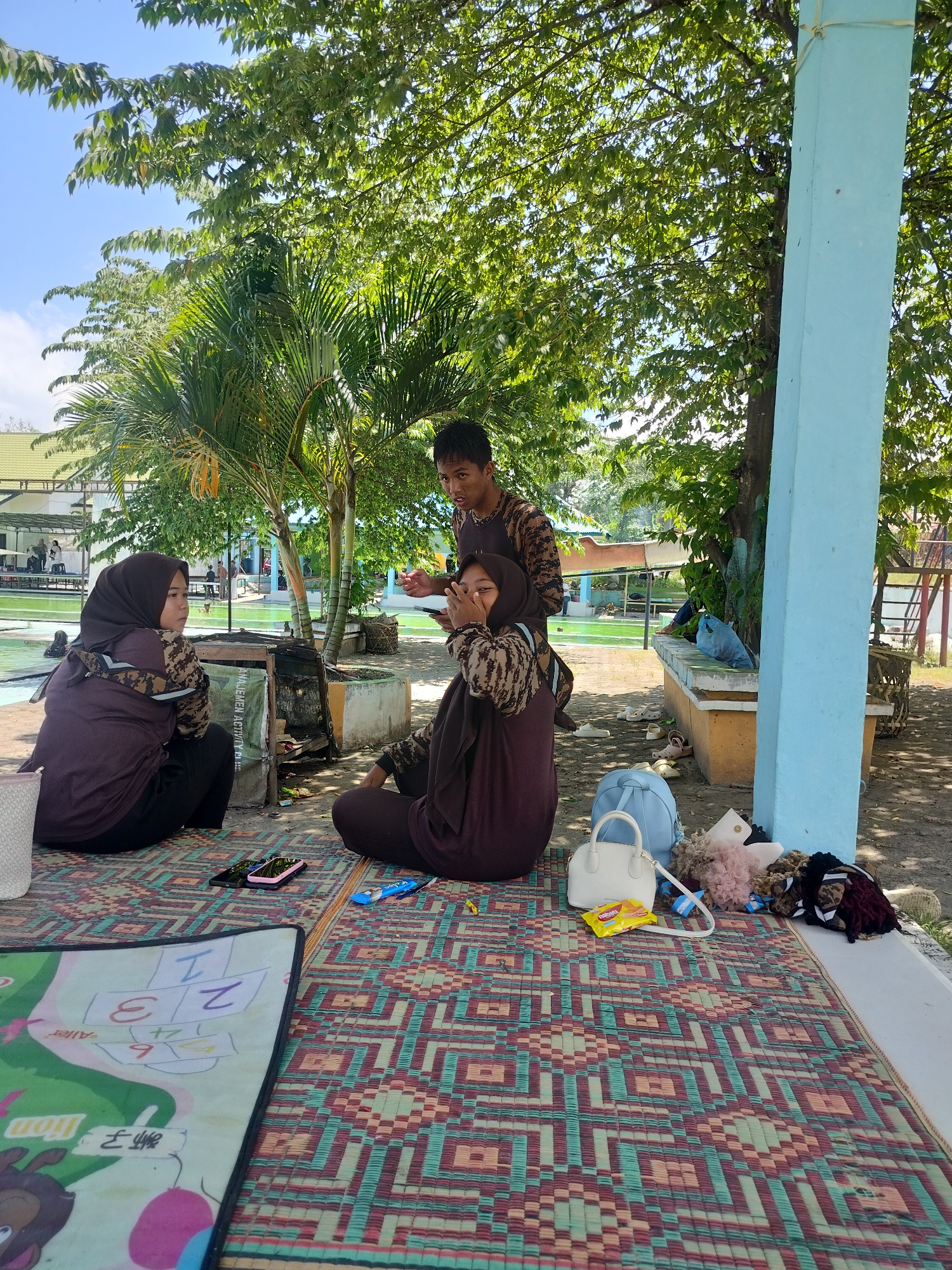 In Tegarmiko, Indonesia, on a sunny day, three young people are sitting on woven mats outdoors. A person wearing a brown patterned shirt stands behind two individuals who are seated. One seated person wears a dark brown hijab and a matching shirt, looking away from the camera. The other seated person, also in a dark brown hijab and shirt, is looking at the camera and covering their mouth and nose with their hand. They are all on a patterned mat. Several items are scattered around them, including a white handbag, a blue backpack, a packet of snacks, a small pink object, and other fuzzy items. A mat with numbers and drawings is visible in the foreground. In the background, palm trees and leafy trees are prominent. Beyond the trees, a building with a green roof and a swimming pool area are visible, suggesting a public space or recreational facility. A light blue pillar stands to the right of the frame.