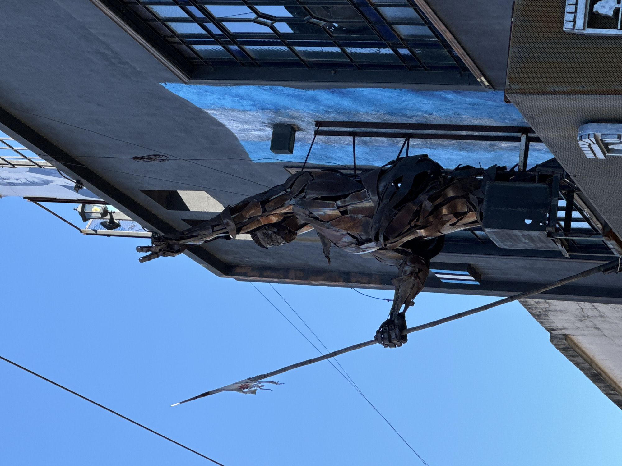 The photo depicts a sculpture of a man holding a spear erected on top of a building in Buenos Aires, Argentina. The sculpture is constructed from metal sheets welded together. The man is in a lunge position, poised to throw the spear. The building has windows and several horizontal blue and white stripes painted beneath it. The sky is clear blue.