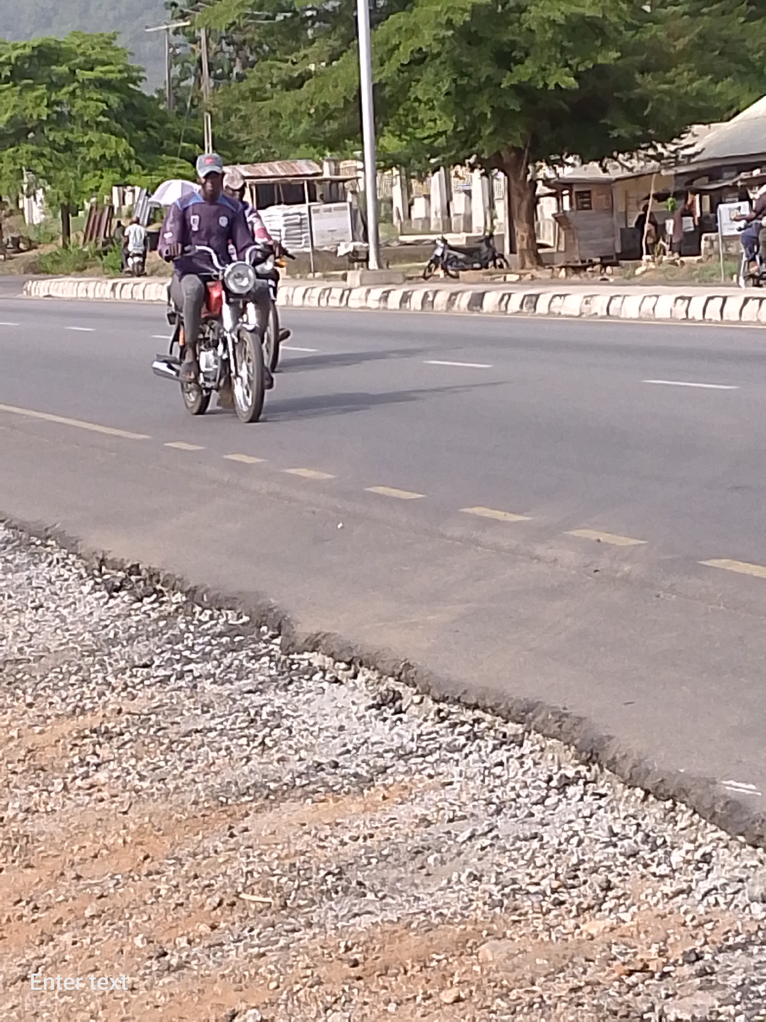 A medium shot depicts two people on a red motorcycle traveling on a paved road in Adankolo, Nigeria. The road features intermittent yellow lines and a concrete curb separates it from a gravelly area on the right side. On the opposite side of the road, there are several small buildings and trees.