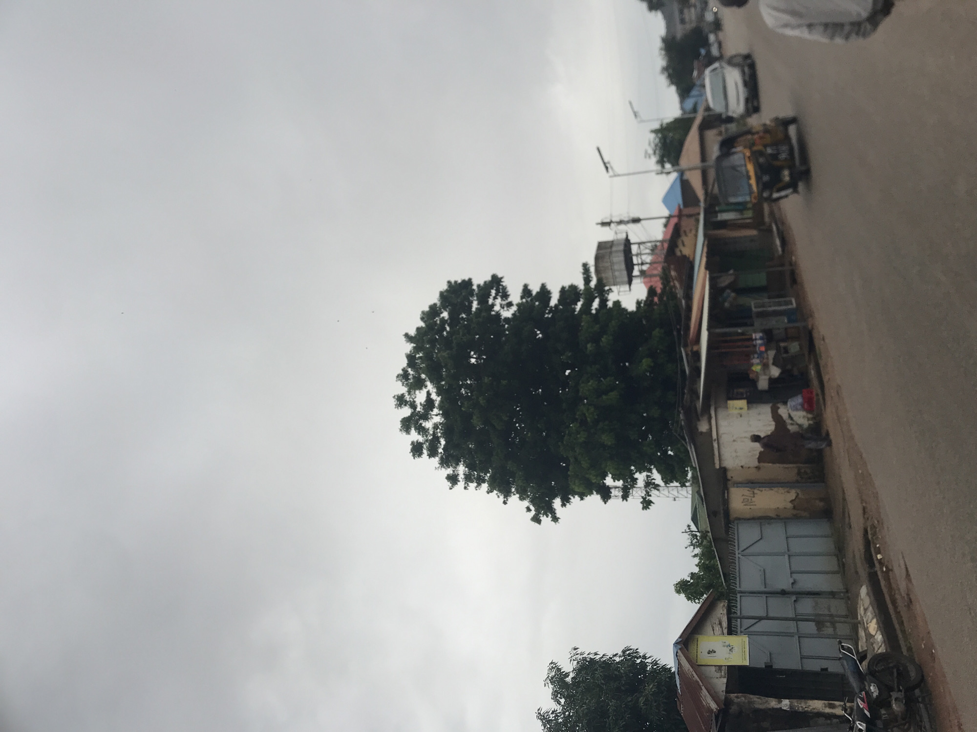 The image shows a street scene in New Stadium, Nigeria, under a cloudy sky. On the left side of the road, there is a metal gate leading to a compound with several small structures. A motorcycle is parked on the left shoulder of the road. Across the street, a large tree stands prominently, with a water tank and a tall communication tower visible behind it. Several small shops are lined up along the right side of the road, with awnings providing shade. A tuk-tuk is in motion on the road, followed by a white car. A person in a grey jacket is walking away from the camera on the right side of the road. The overall impression is of a busy, functional urban environment.