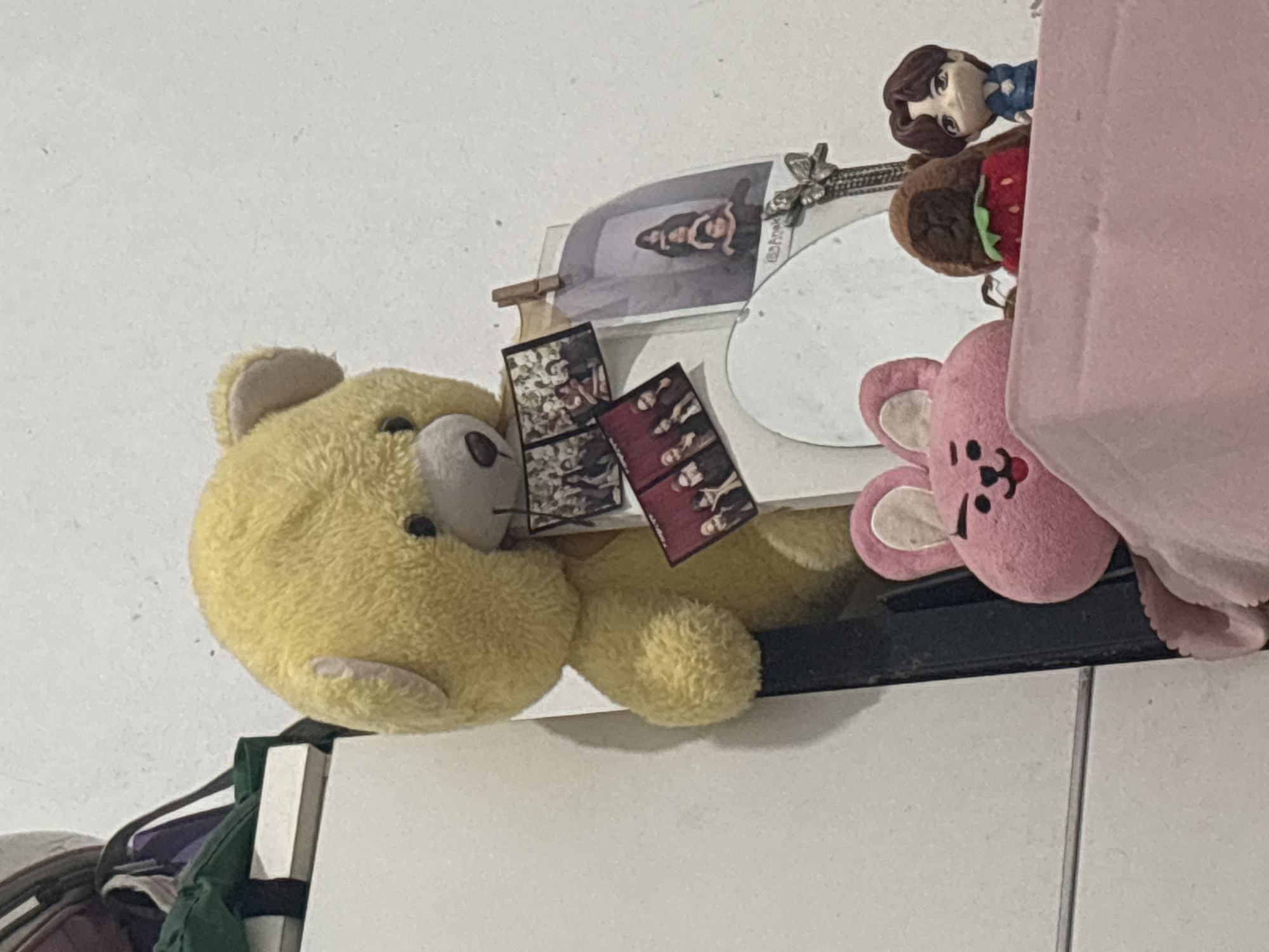 The image shows a collection of plush toys and figurines arranged on a shelf against a white wall. In the foreground, a pink plush bunny-like toy with black eyes and a pink tongue is partially visible. To its right, a small brown plush toy with a red strawberry on its chest and a small blue figurine with dark hair are placed. Behind these, a large yellow teddy bear is positioned, holding several small photos clipped to its paws. One of these photos shows four people standing in a formation, while another displays a group of individuals. A larger, framed picture hangs from a clip on the wall behind the teddy bear, also featuring a group of four people. The overall setting appears to be a domestic indoor environment. The specific location is identified as North Jakarta, Indonesia.