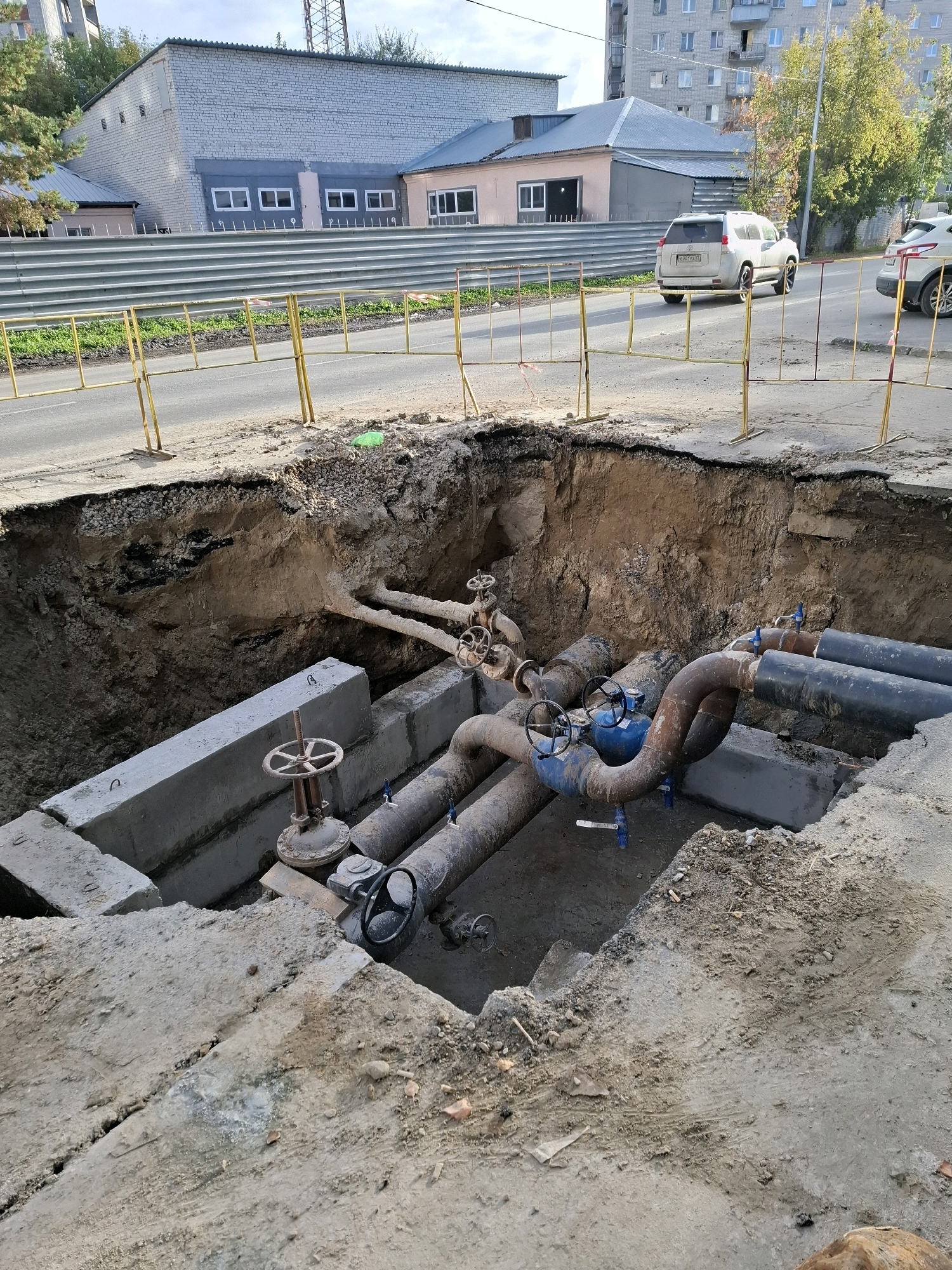 A construction site in Tyumen, Russia, features a large, excavated pit on the side of a road. The pit exposes a network of large pipes and valves, suggesting utility work. Yellow construction barriers and fencing line the road, which has some visible vehicle tracks. In the background, a brick building and a multi-story apartment building are visible. The sky is partly cloudy.