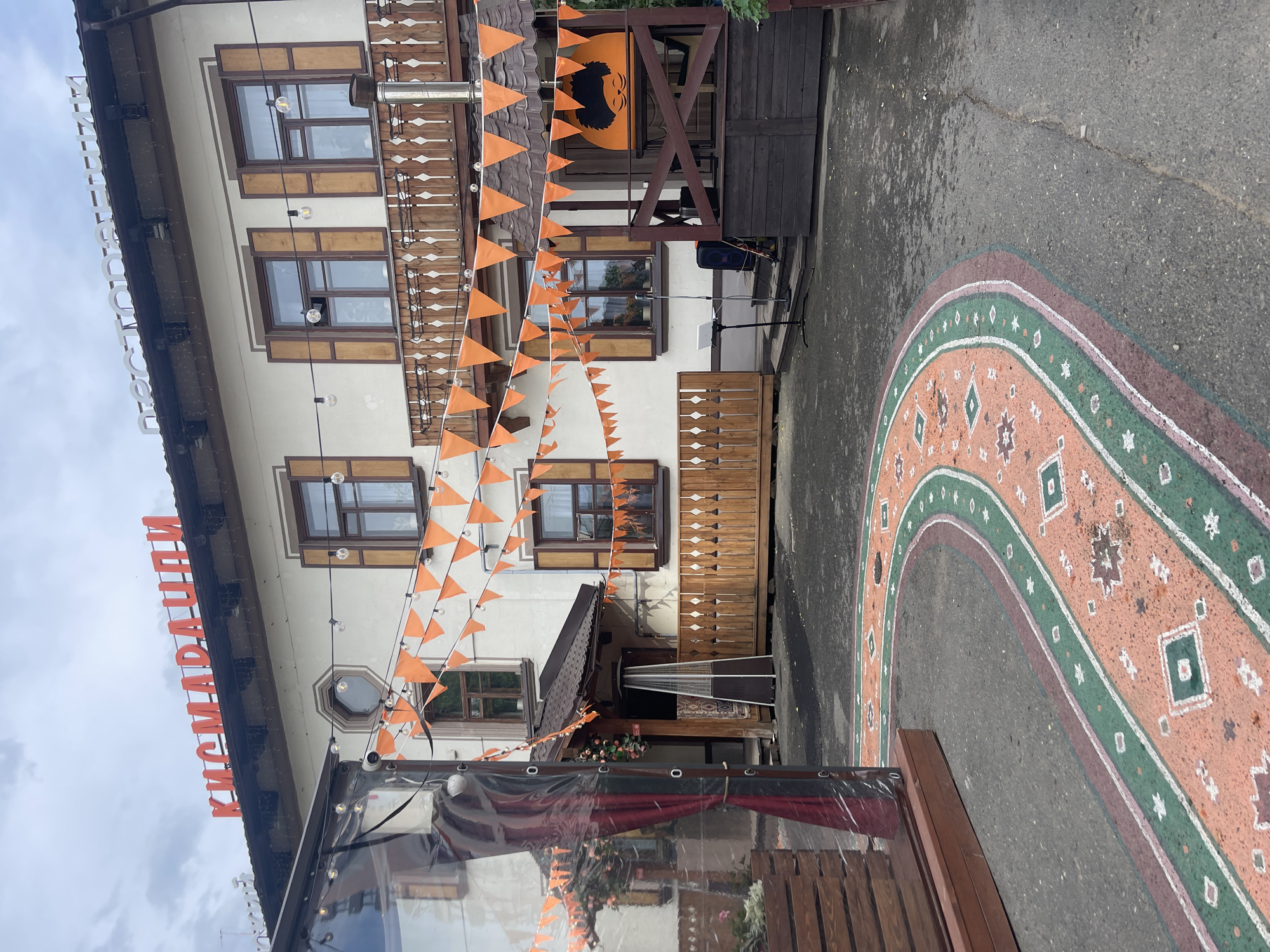 The image shows the exterior of a building in Perm, Russia. The building has a cream-colored facade with brown wooden window frames and shutters. Orange pennant banners are strung across the front of the building. A large sign with red Cyrillic letters is mounted on the roof. A portion of a painted rug pattern is visible on the pavement. The sky is overcast.