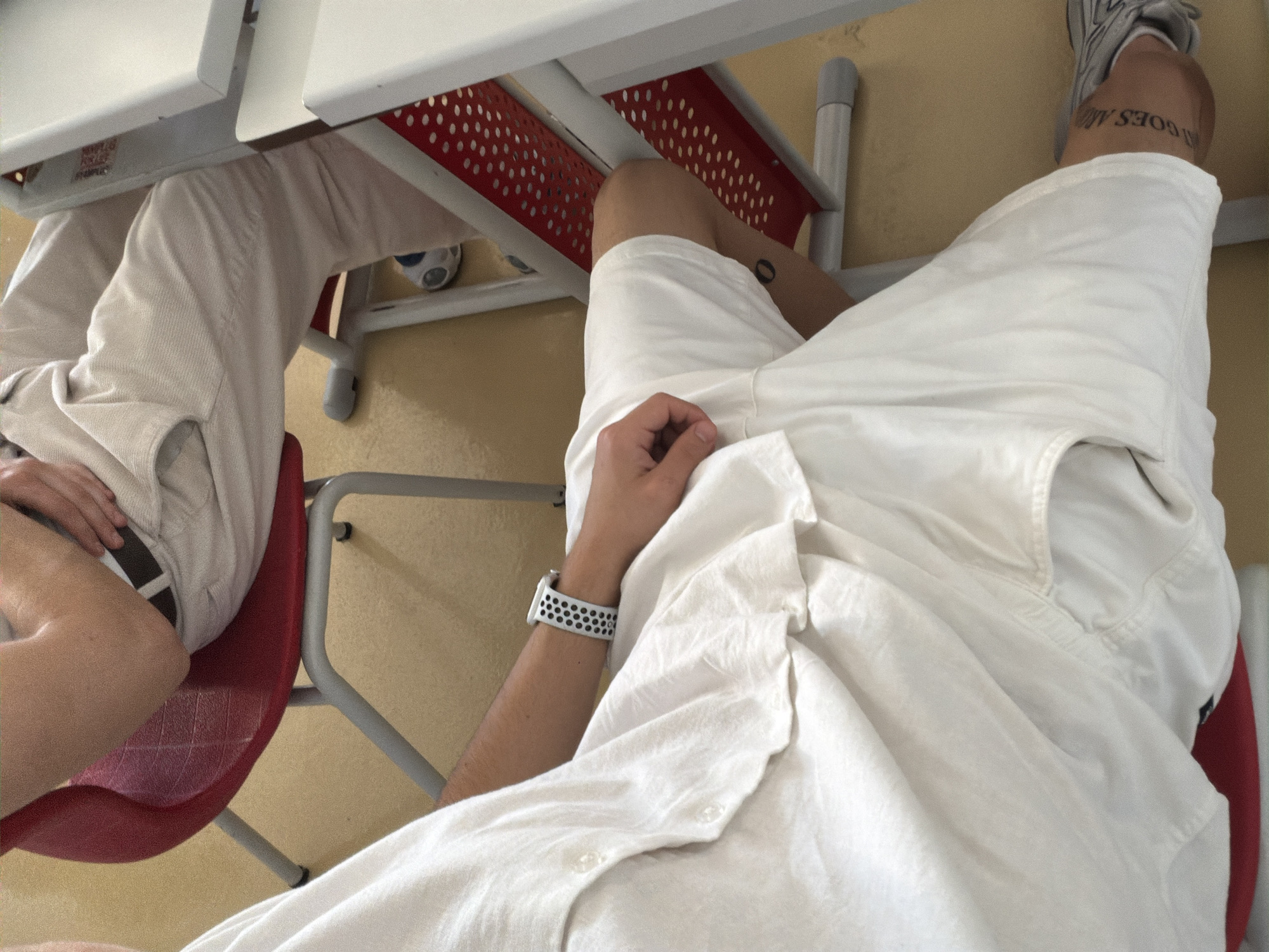 The image depicts an indoor scene, likely a classroom or lecture hall, with two individuals seated at desks. The floor is a light brown linoleum. In the foreground, a person wearing a white linen shirt and white shorts is seated on a red chair. The person has a white and black patterned watch on their left wrist. A tattoo is visible on their right thigh, with the visible portion reading "GOES AM."
In the background, another person is seated on a similar red chair, wearing light-colored pants and a dark belt. Part of a white desk with a red perforated metal support is visible to the right. The lighting suggests an artificial light source, likely overhead. The image does not contain any specific visual cues that would allow for its location to be identified as Plovdiv, Bulgaria.