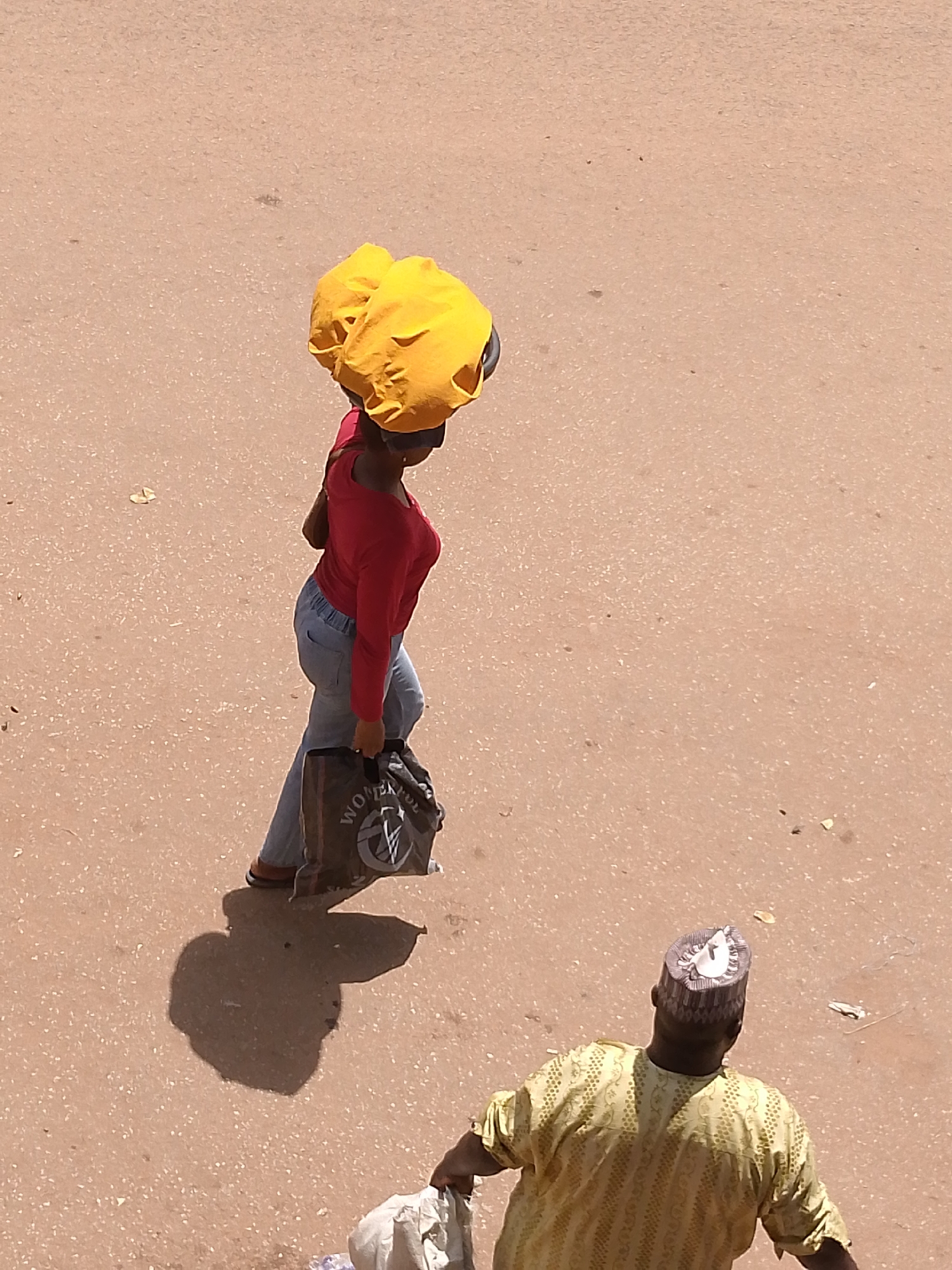 An overhead shot shows a woman in a red long-sleeved shirt, light blue jeans, and sandals carrying a dark bag in her right hand and a large yellow bundle on her head. Her shadow is visible on the ground to her left. To the lower right, a man in a yellow and white shirt and a small patterned white and brown cap walks with a plastic bag in his left hand. The ground is tan with small dark spots, and light debris is scattered around the people. This scene is located in Bukuru, Nigeria.