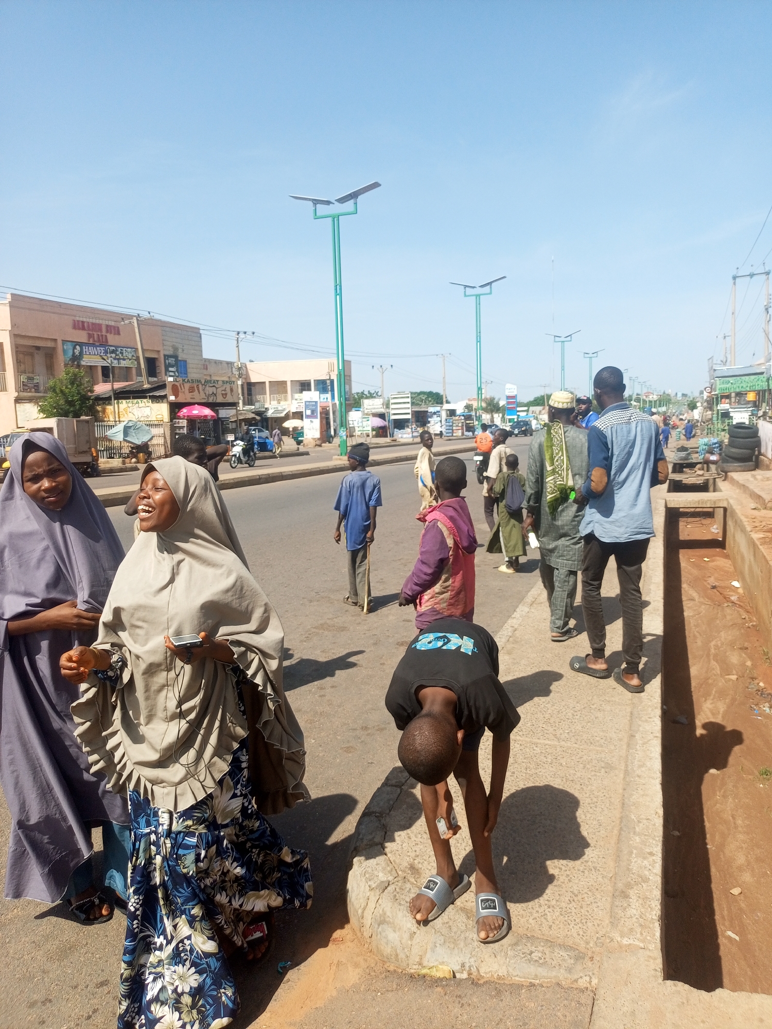 The image depicts a street scene in Tempure, Nigeria, likely during daytime given the bright sunlight casting distinct shadows. The setting is a paved road with buildings and shops lining one side. Several individuals, including men, women, and children, are present on the street and sidewalk.
In the foreground, two women wearing hijabs are engaged in conversation. One woman, on the left, wears a dark purple hijab. The woman on the right, who is laughing or speaking animatedly, wears a lighter grey hijab and a colorful floral patterned skirt. She is holding a mobile phone. To the right of the women, a young boy in a black t-shirt and grey sandals is bent over, appearing to be picking something up from the ground or tying his shoe. Further down the street, other individuals are visible walking, talking, or standing. A man in a blue shirt and dark pants walks away from the camera, while other people are seen in the background near shops and along the road.
The environment suggests a typical urban or semi-urban street with ongoing daily activities. Streetlights with solar panels are visible along the road, indicating infrastructure development. Shops with signs are present, though the text on most is not clearly legible. One sign reads "ALHAMDU LILLA" and another appears to be "HAWEE'S". The overall atmosphere seems casual and bustling, with people going about their day under a clear, bright sky.