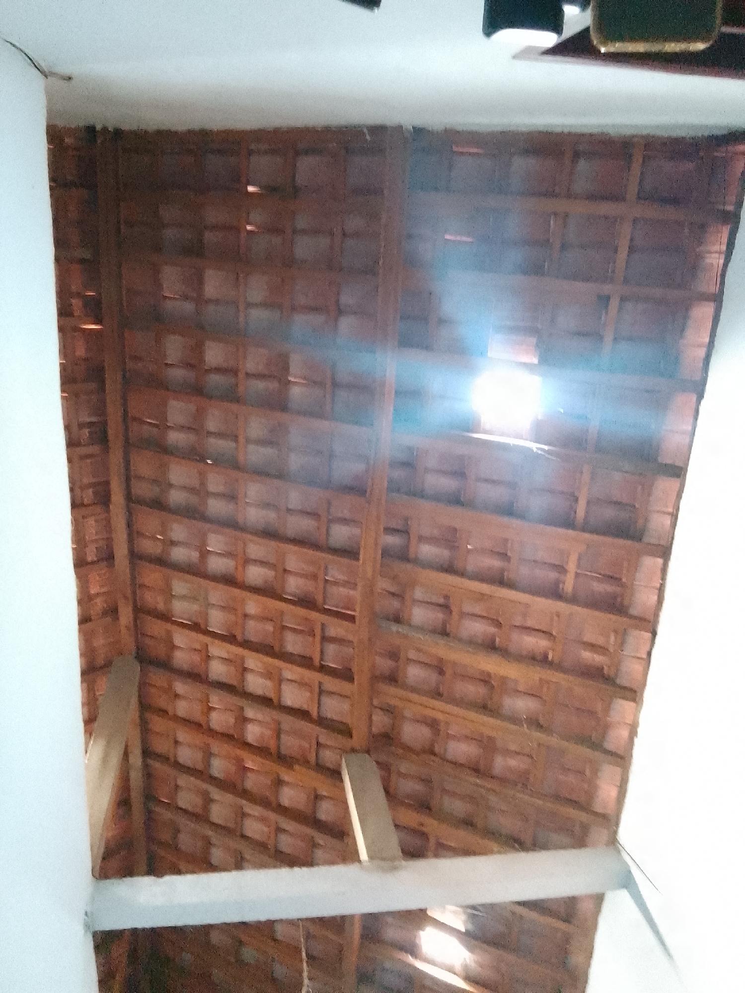 The image shows a close-up view of a tiled roof from below. The roof is constructed with reddish-brown ceramic tiles arranged in a grid pattern, supported by wooden beams and rafters. The lighting suggests it's daytime, with sunlight filtering through gaps in the tiles, creating bright spots and lens flares. On the left and right edges, parts of white walls are visible, indicating an interior space beneath the roof. The context is a residential or commercial building in Karangan, Indonesia, likely a traditional architectural style.