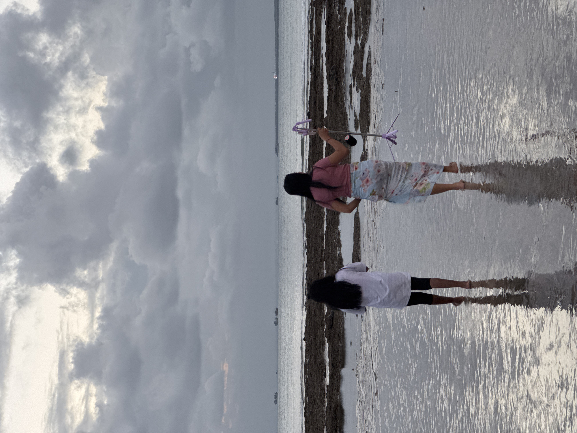 The image depicts two young females on a wet, sandy beach, with the ocean in the background. One girl, in the foreground, wears a white t-shirt and black shorts, standing with her feet in shallow water, her reflection visible on the wet sand. Behind her, another girl is partially visible, wearing a pink t-shirt and a floral skirt, holding a purple object on a stick.
The scene is set at what appears to be low tide, with dark, rocky formations jutting out from the water. The sky is overcast with dramatic, grey clouds, suggesting a storm might be approaching or has recently passed. The lighting is dim, indicating it is likely late afternoon or early evening. The ocean stretches out to the horizon, where a few distant boats or ships are visible.
The overall mood is calm and serene, despite the cloudy weather. The reflections on the wet sand add a reflective and slightly surreal quality to the image. The location, Kuta Selatan, Indonesia, is known for its beaches, and this scene captures a quiet moment by the sea. No visible text is present in the image.