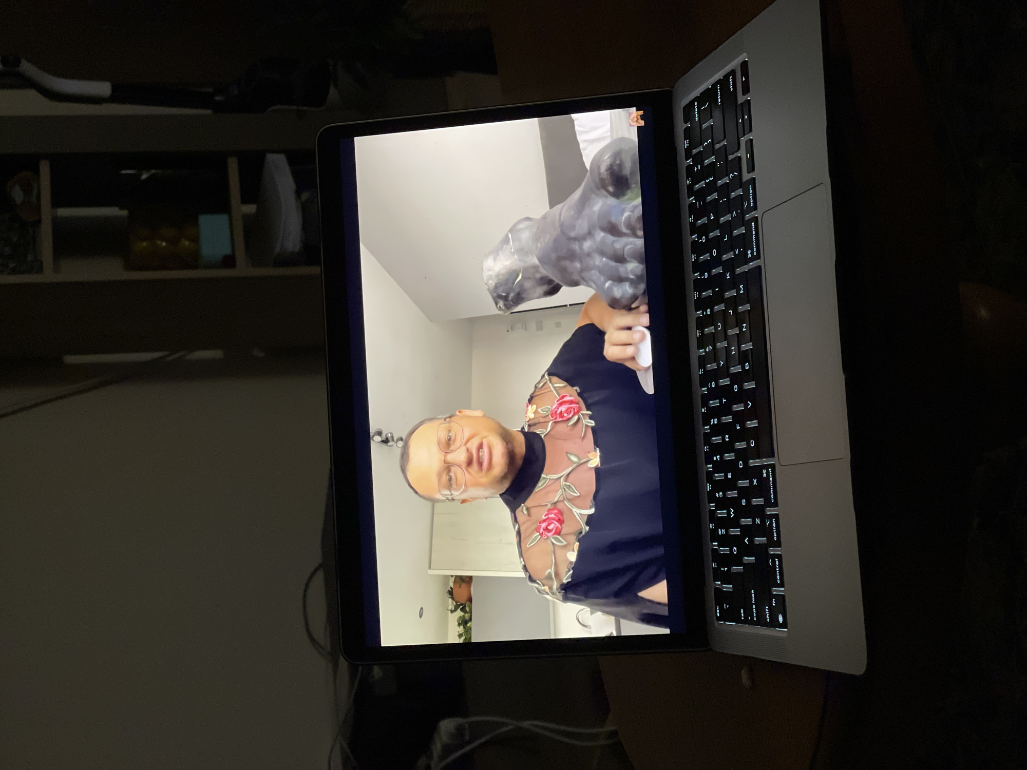 The image shows an open laptop displaying a video call. The video features a person holding a grey sculpture. The person has short hair, wears glasses, and is wearing a black top with floral embellishments. The background of the video is a white wall with some visible kitchen elements. The laptop is resting on a dark surface. There is a shelf visible in the background behind the laptop. The location cannot be inferred from the image.
