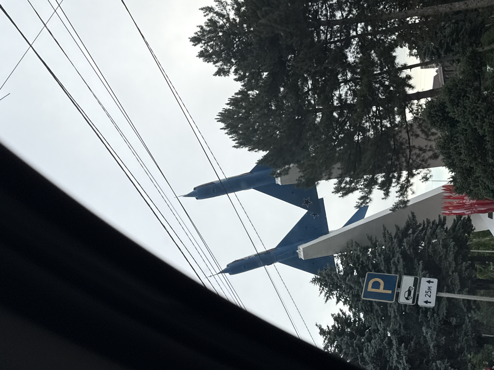 A low-angle, outdoor, eye-level shot captures a monument consisting of two blue fighter jets angled upwards. The jets are positioned on a white, angled pedestal against a pale, overcast sky. A dark, curved object, possibly a car window frame, partially obscures the left side of the image. In the foreground, several tall, dark green evergreen trees frame the monument and obscure the lower portion of the scene. A traffic signpost with three signs is visible to the left of the monument. The top sign is a blue square with a white "P" for parking. The middle sign is a white rectangle with a black silhouette of a car. The bottom sign is a white rectangle with a black upward-pointing arrow and "25M" indicating a distance. Red flags are visible in the background to the right, draped along a low wall or structure. The overall impression is that of a memorial or historical monument.