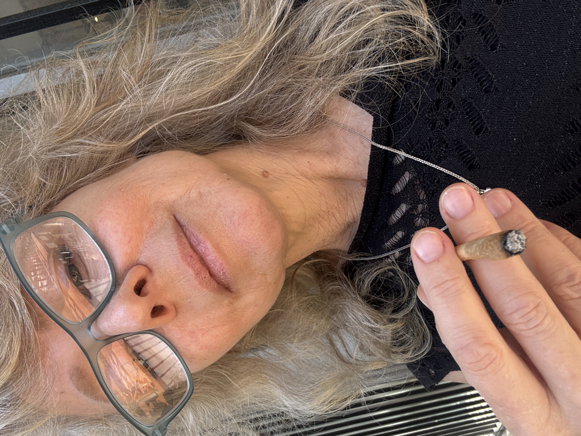 A woman with gray hair and glasses lies on her side, holding a lit marijuana cigarette (joint) in her right hand. She is wearing a black, lace-like top and a silver necklace. The scene is likely indoors. The reflection in her glasses suggests an urban setting with buildings, possibly in Tel-Aviv, Israel. The background contains striped lines, potentially part of a furniture or architectural element.