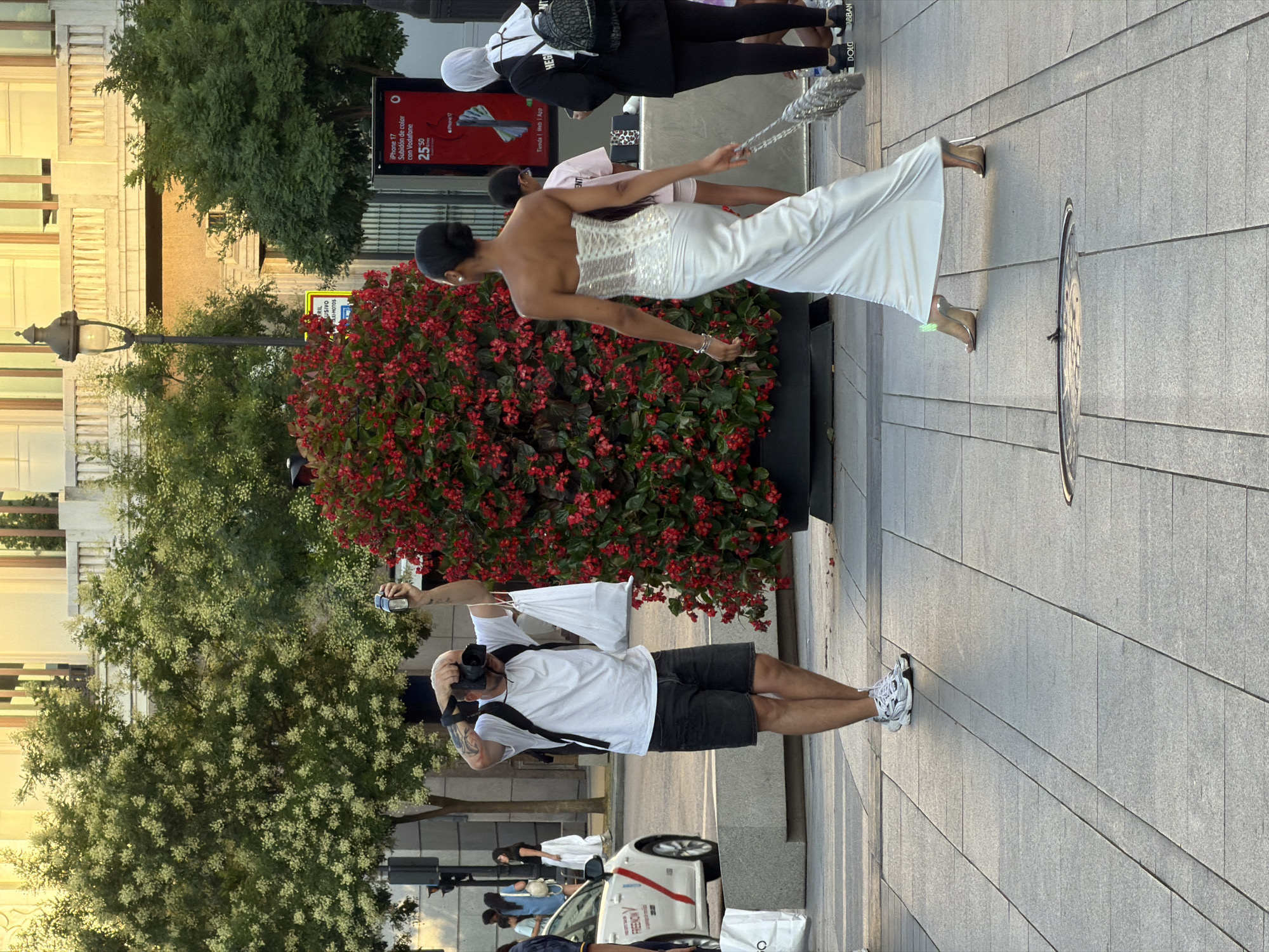 In Madrid, Spain, a photographer, wearing a white t-shirt, black shorts, and white sneakers, stands on a paved walkway, holding a camera up to take a picture. He has a backpack on and is carrying a white bag in his left hand. To his right, a woman in a long, white, strapless dress with silver embellishments walks away from the camera, carrying a silver mesh bag. Behind them, a large planter filled with red flowers is visible, with trees and buildings in the background. Several other people are also present, including a person in a white headscarf to the right. A white taxi is parked on the left side of the image.
