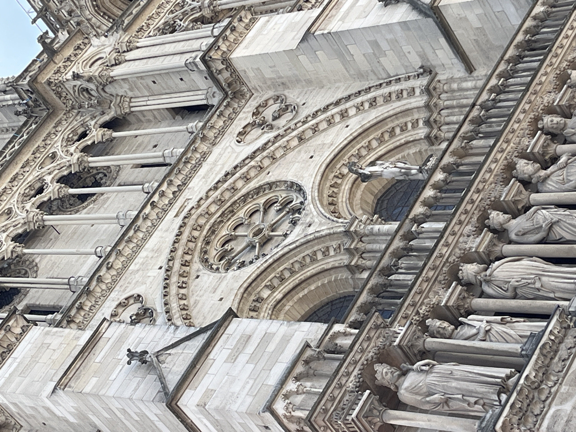 The image shows a close-up, low-angle view of the facade of Notre Dame Cathedral in Paris, France. The architecture is Gothic, with intricate stone carvings, pointed arches, and decorative detailing. Several statues of robed figures, likely kings or saints, are visible in niches along the lower part of the facade. Above them, a rose window with intricate tracery is partially visible, and further up, more architectural ornamentation and columns can be seen. Gargoyles are also present on the upper ledges. The sky is a pale, overcast blue. The lighting is diffused, suggesting it is daytime. The stone of the cathedral is weathered and pale, indicating its age. The image focuses on the details of the construction and ornamentation, highlighting the craftsmanship of the cathedral.