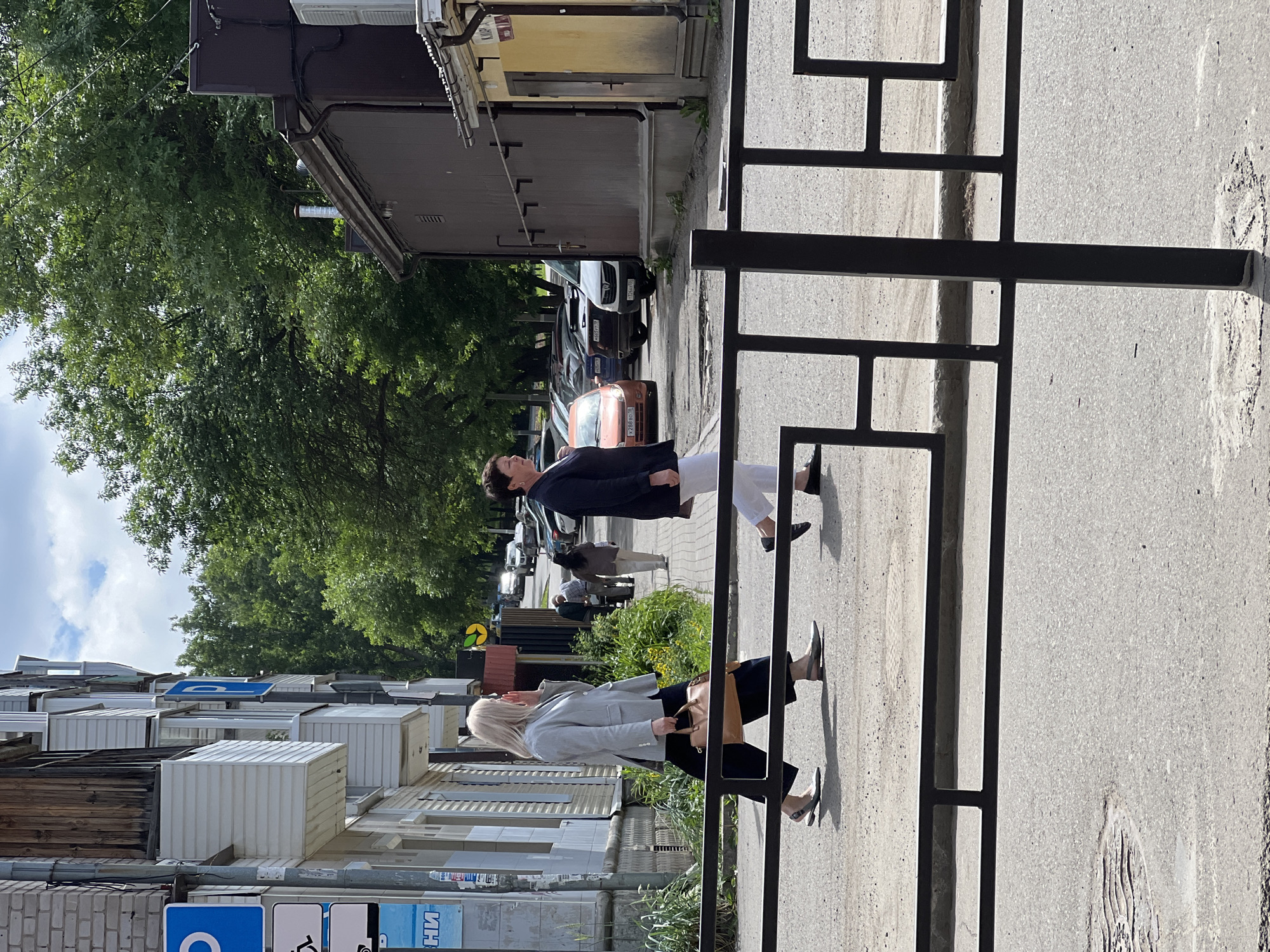 In Izhevsk, Russia, on a sunny day, two women are crossing a paved street near a building with a brown facade. The woman on the left, with blonde hair, wears a grey blazer, dark trousers, and is carrying a light brown handbag. The woman on the right, with dark hair, is dressed in a dark blazer and white trousers. In the background, several cars are parked along the street, and lush green trees line the area. Buildings with balconies and signs are visible on the left side of the image. A black metal fence runs across the foreground, partially obscuring the view of the street and pavement.