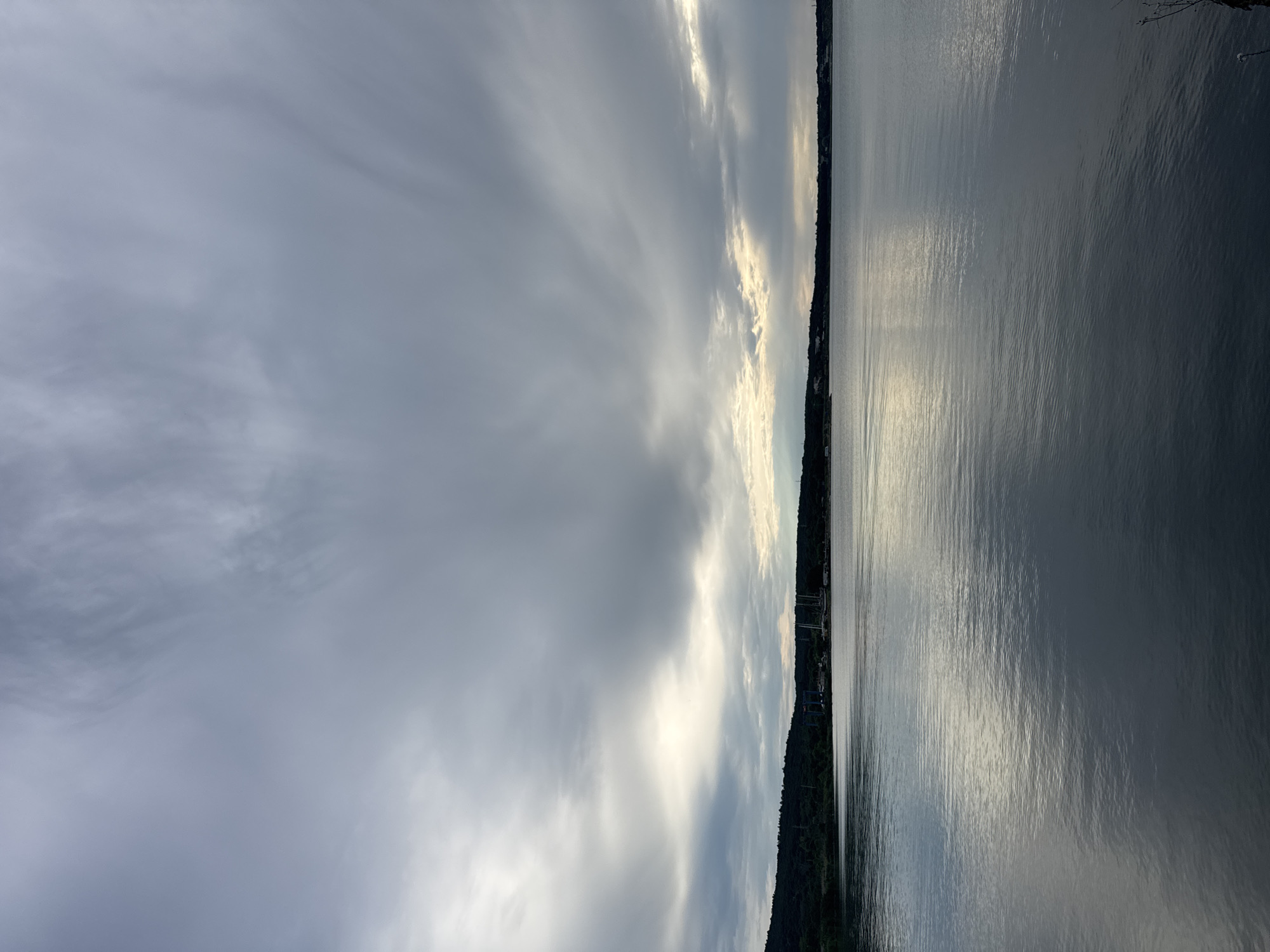 The image depicts a body of water under a cloudy sky, with a tree line along the distant shore. The sky is filled with grey, wispy clouds, with breaks of brighter light suggesting sunlight filtering through. The water is calm, with subtle ripples reflecting the sky. Along the horizon, a dark, densely treed landscape is visible, dotted with structures. On the far left, a tall, dark blue industrial-looking structure with a gantry is prominent. Further to the right, several tall masts, likely belonging to sailboats, are visible. The overall scene suggests a harbor or a bay area. The location is identified as the Burgas Bay, near the city of Burgas, Bulgaria.